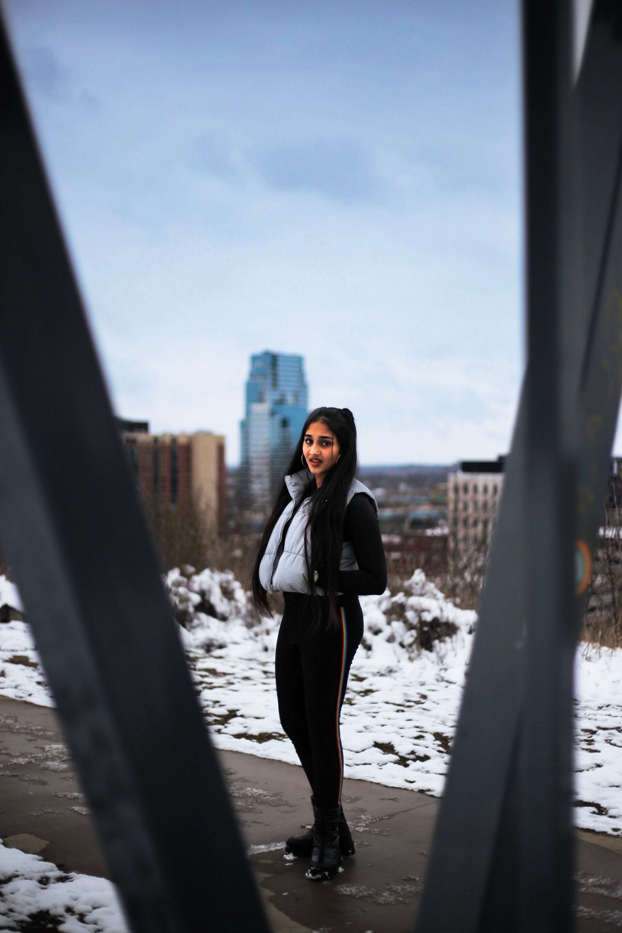 Smiling young woman in silver vest and black outfit poses between metal railings with snowy cityscape backdrop.