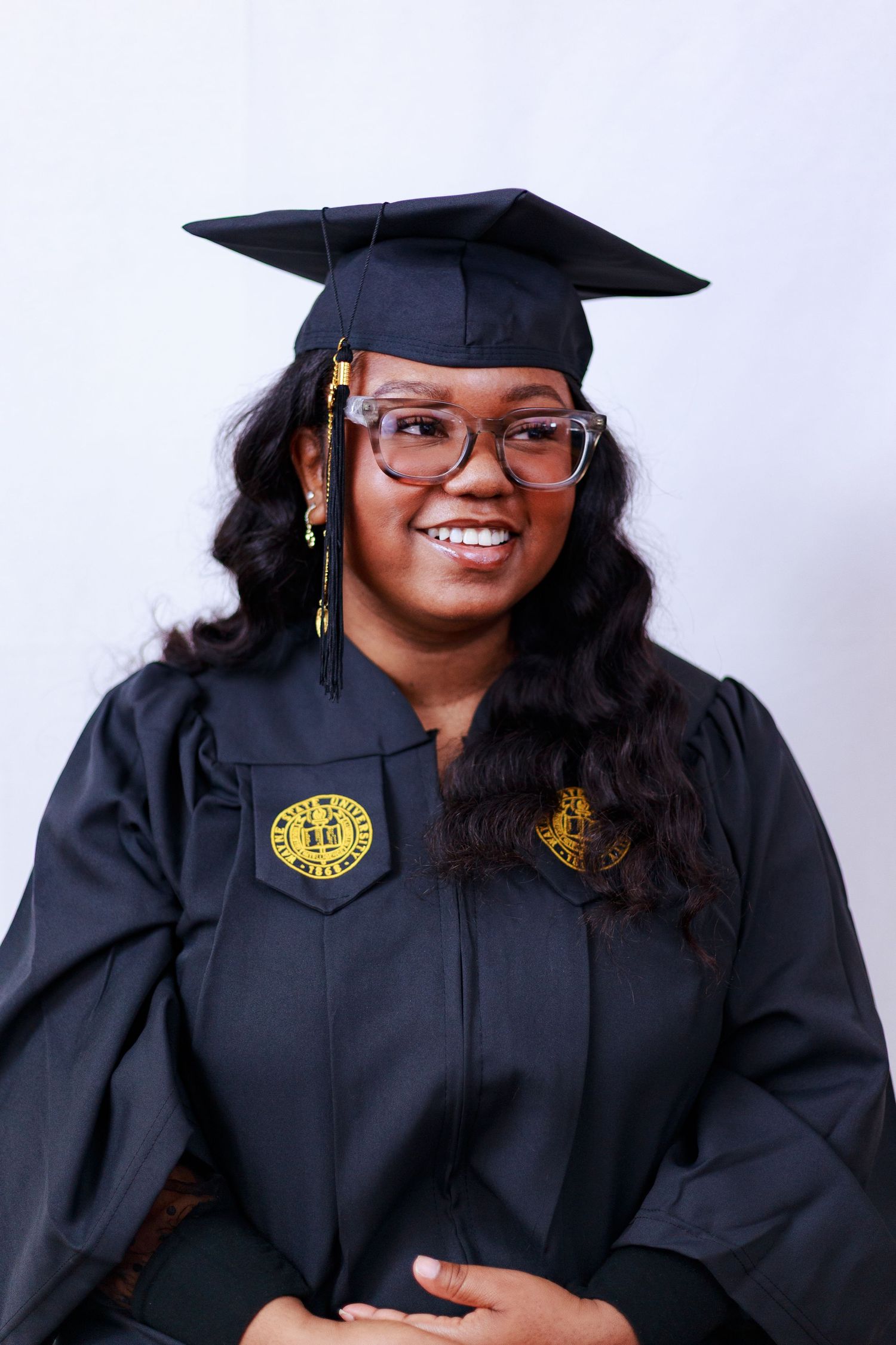Young woman in black graduation cap and gown with gold university emblems smiling during graduation portrait session.