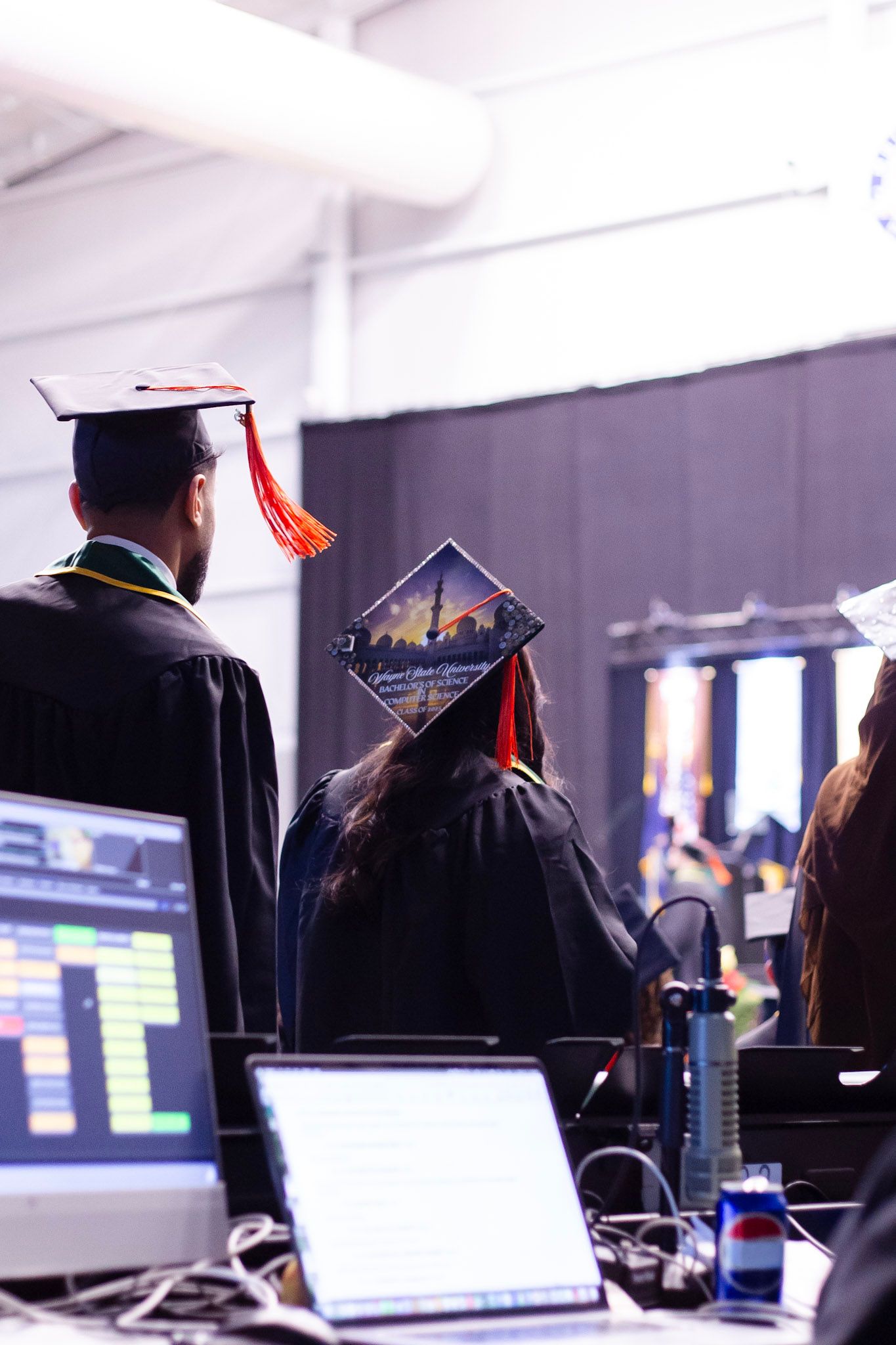 Two graduates in decorated mortarboard caps stand backstage near audio equipment at commencement.