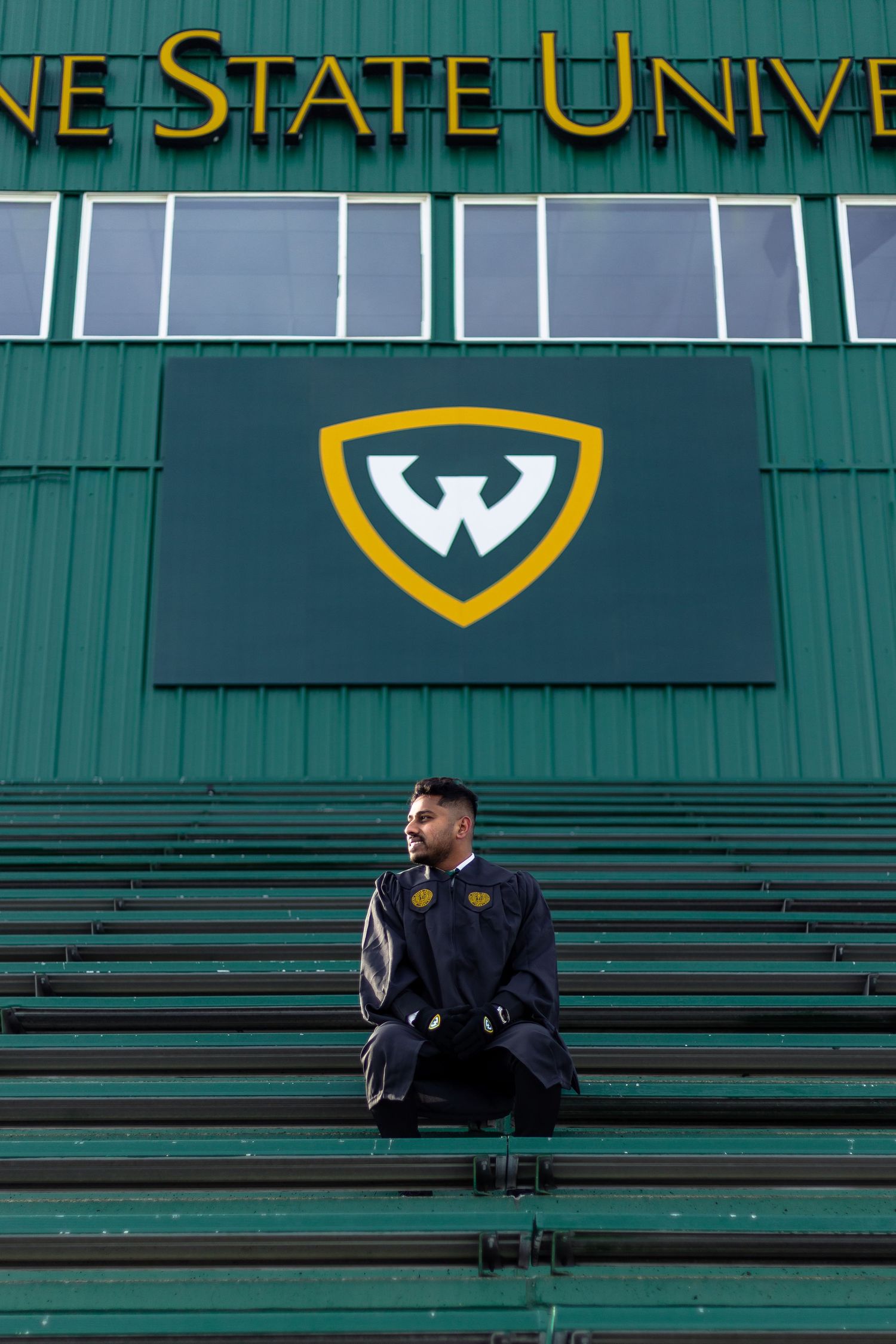 Wayne State University graduate in cap and gown sitting on green stadium bleachers with WSU logo.