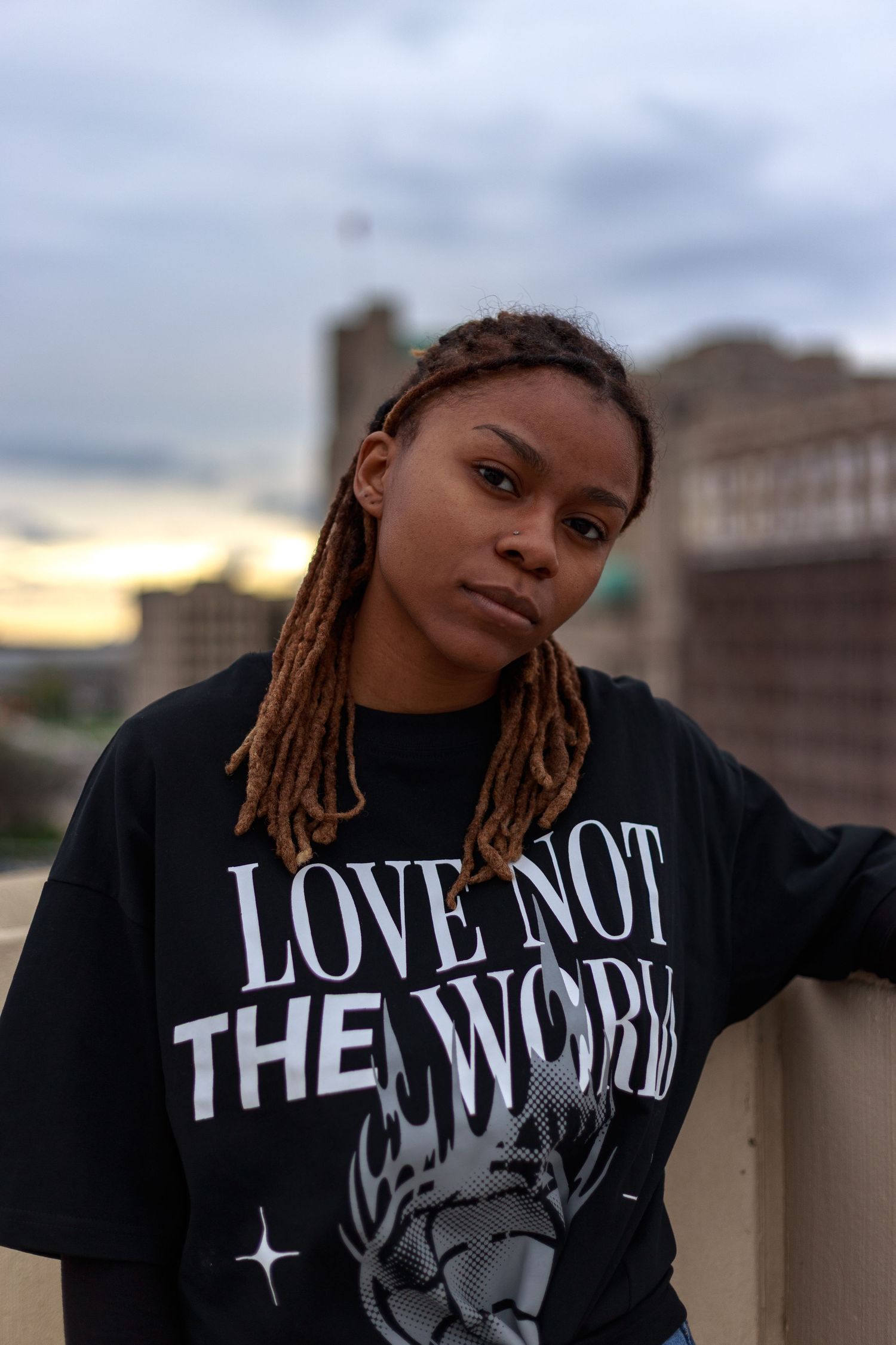 Young woman with locs in an oversized black Love Not The World tee poses on an urban rooftop at dusk.