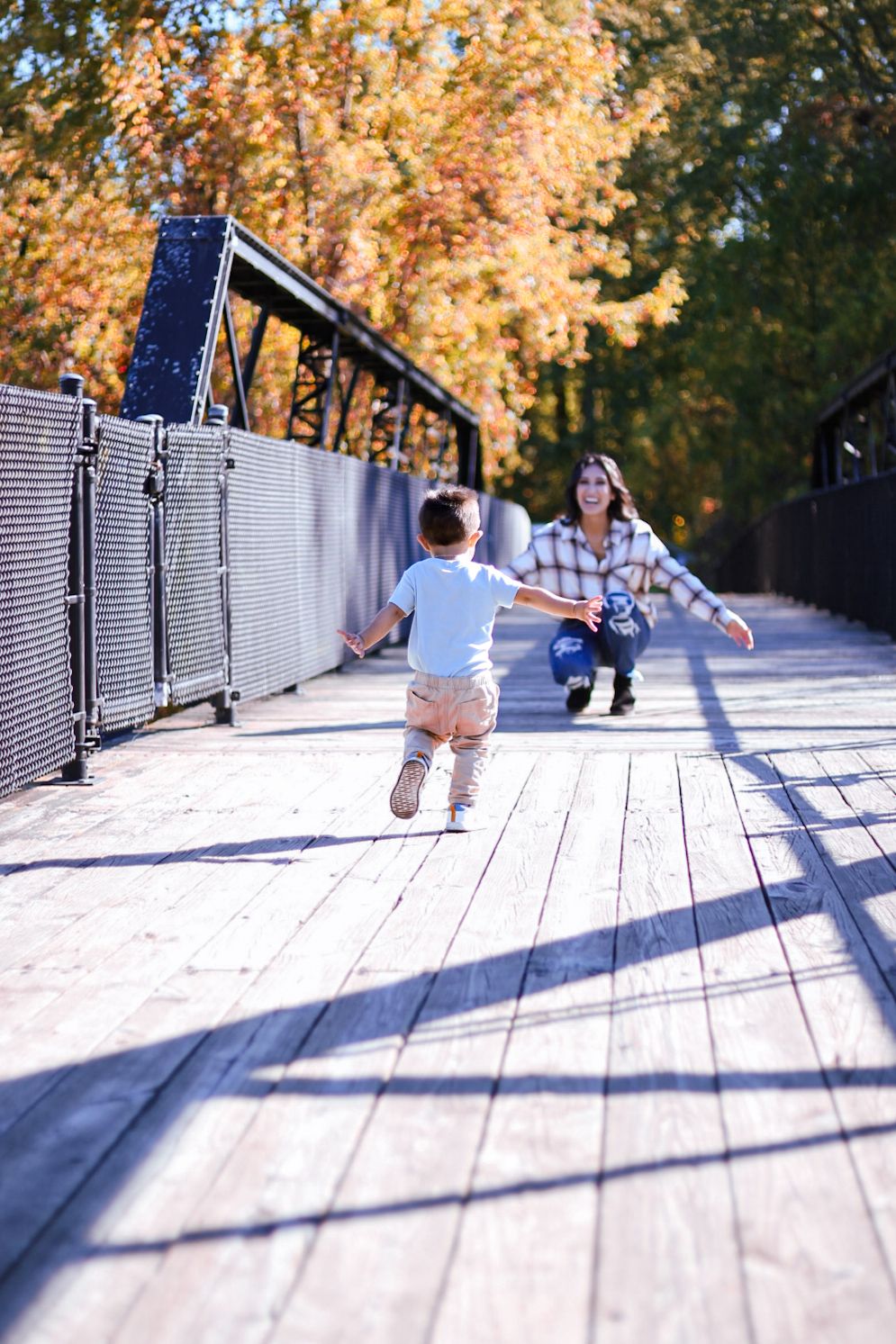 Toddler boy running toward crouching mother on wooden bridge surrounded by golden fall trees.