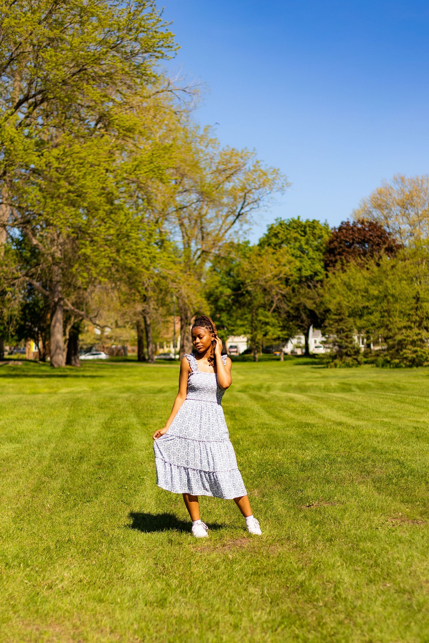 Young Black woman in blue patterned midi dress poses on green lawn in sunny park.