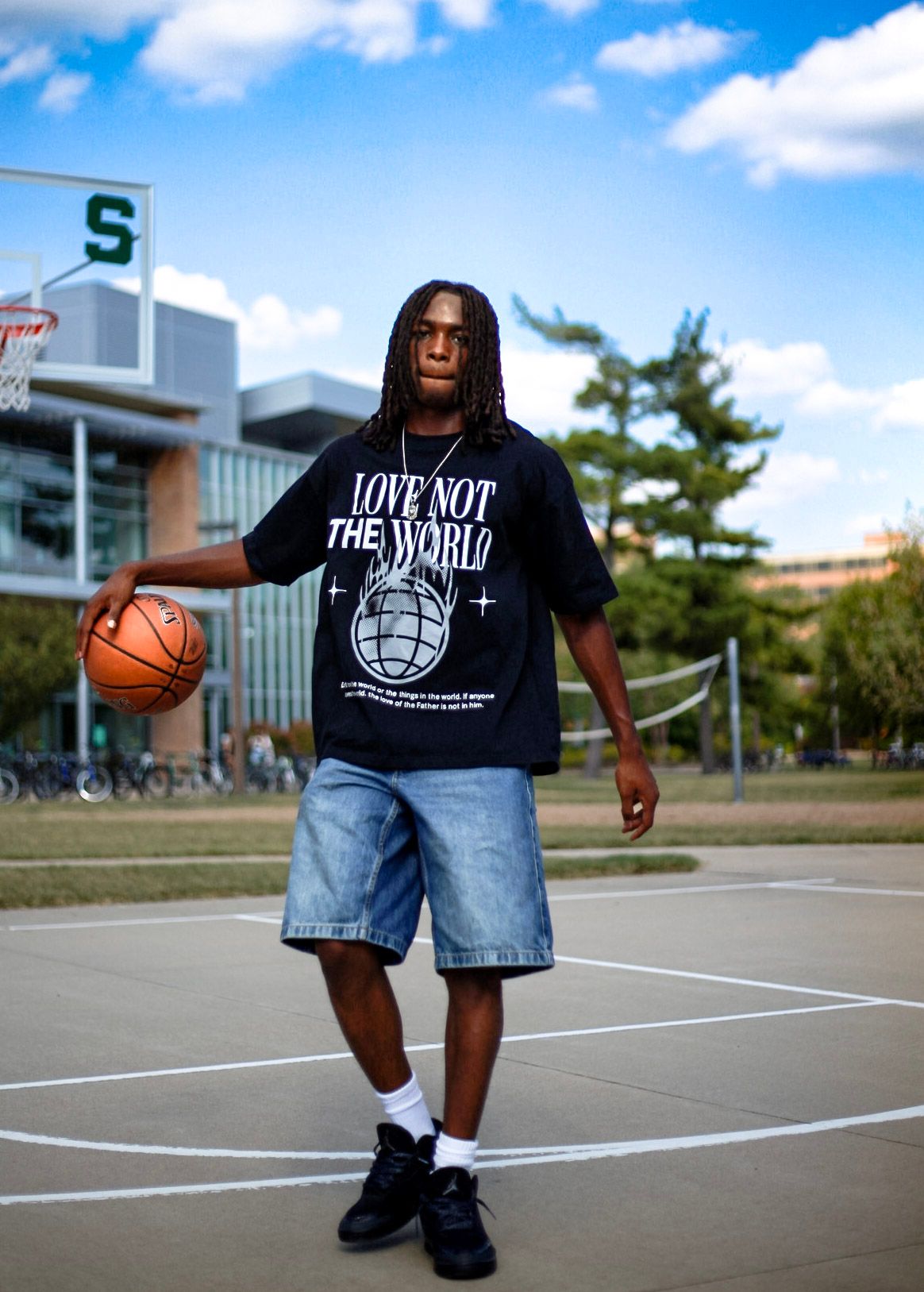 Man holding basketball in black Love Not The World graphic tee on sunny outdoor court.