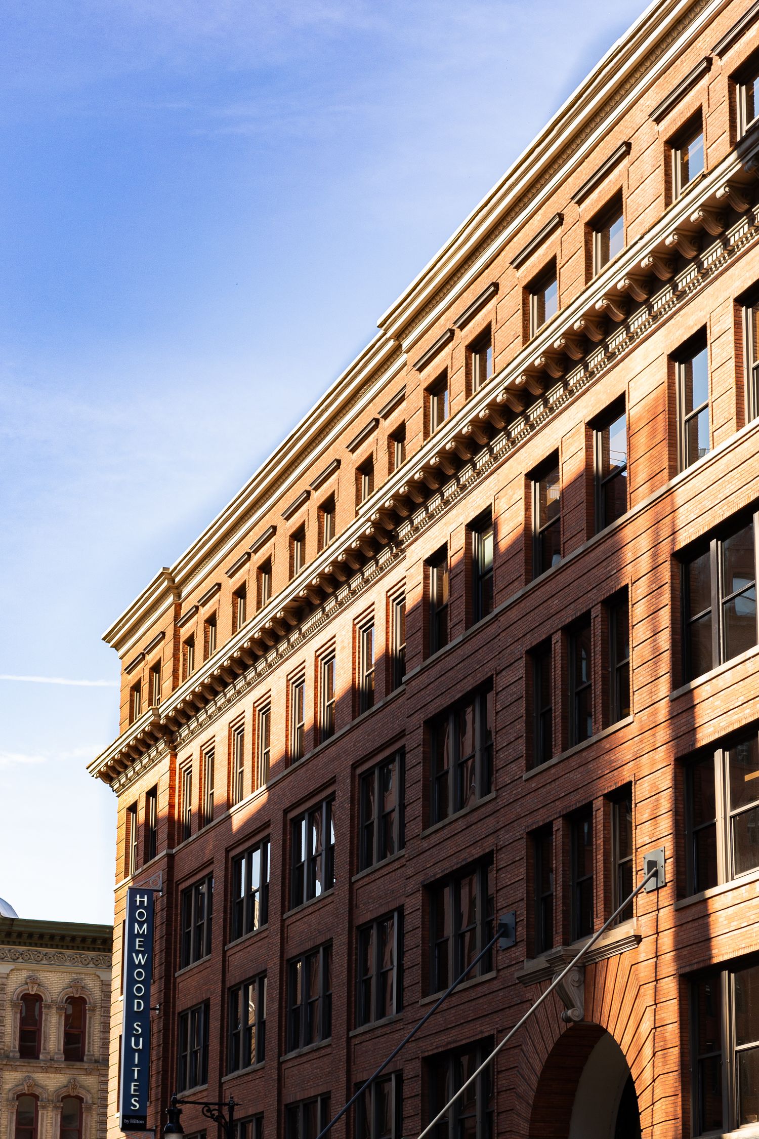 Historic red brick building with ornate cornice details against a blue sky, featuring the Homewood Suites sign.