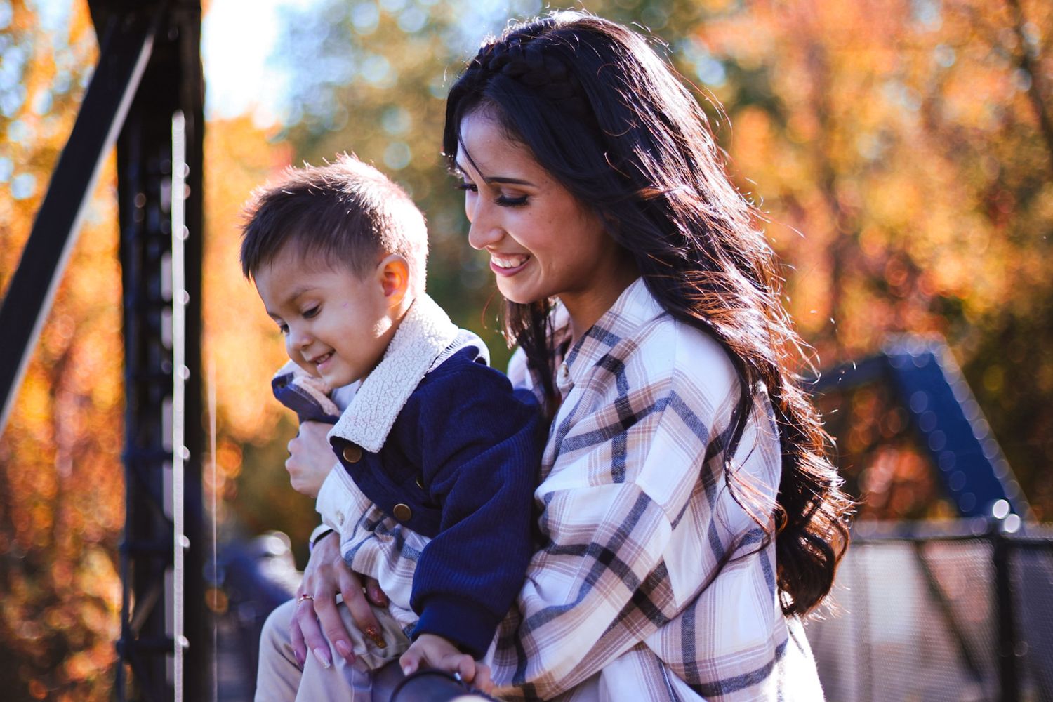 A young mother carries her toddler outdoors amid vibrant orange and yellow autumn leaves.