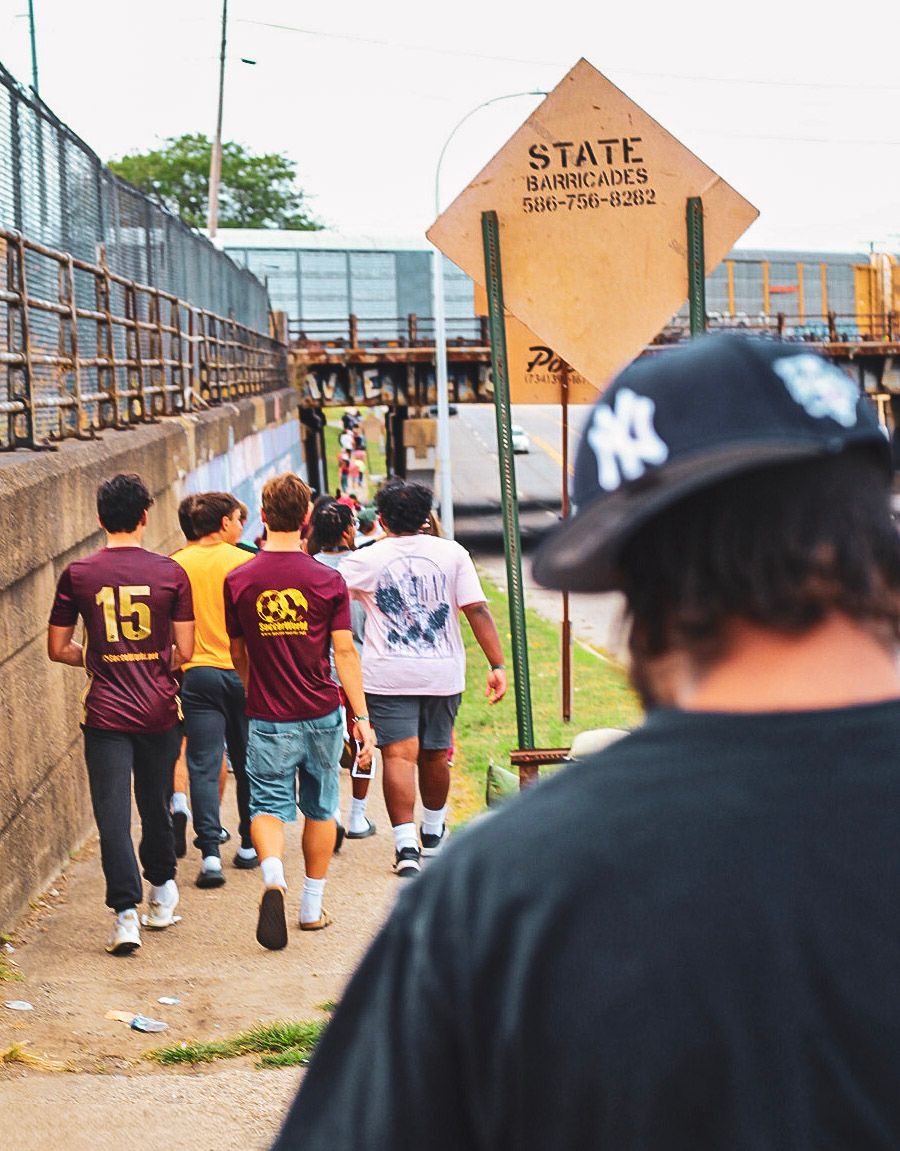 Group of young soccer fans in maroon jerseys walking toward a stadium entrance past a State Barricades sign.