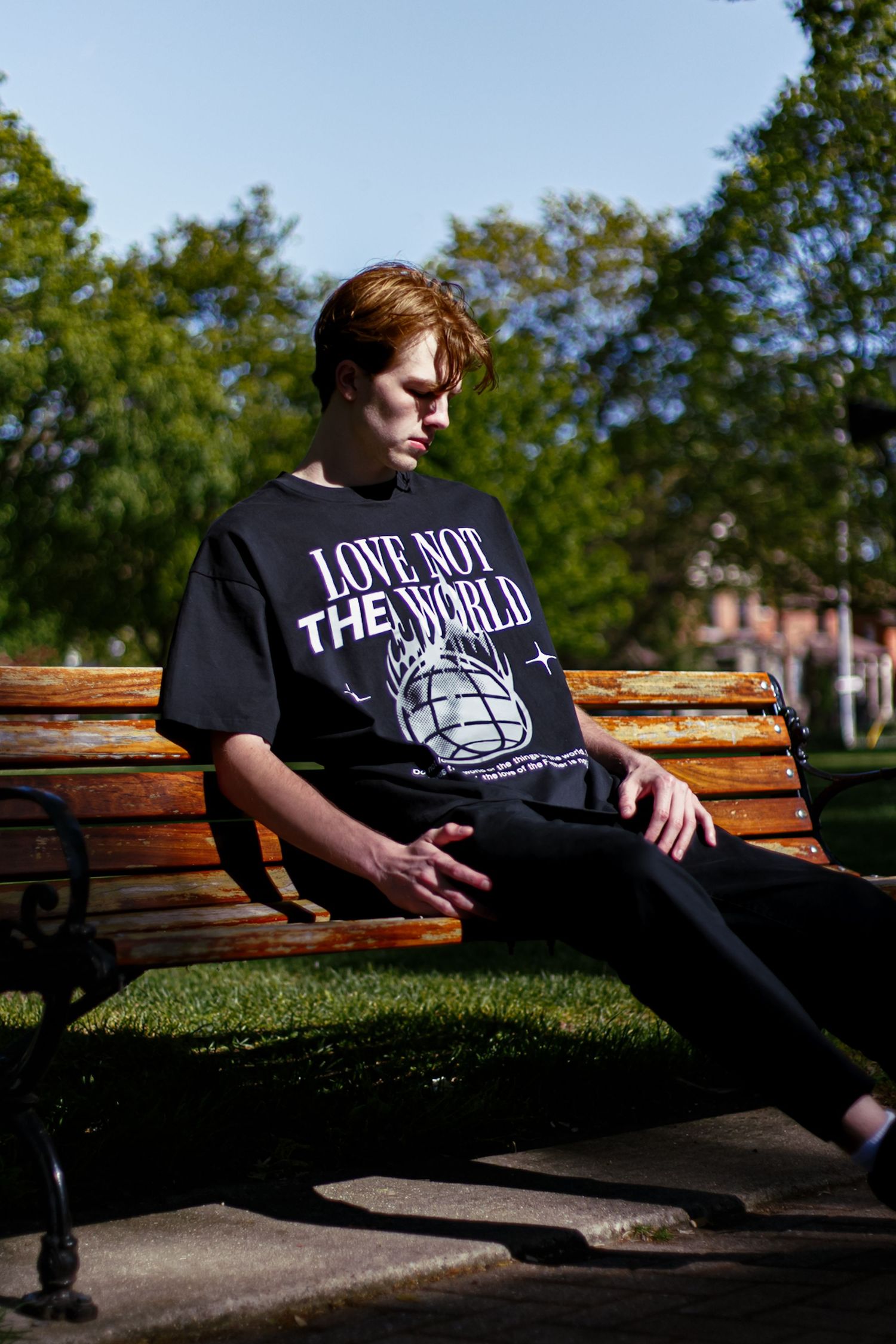 Young man lounging on park bench wearing black Love Not The World graphic tee in sunny park.