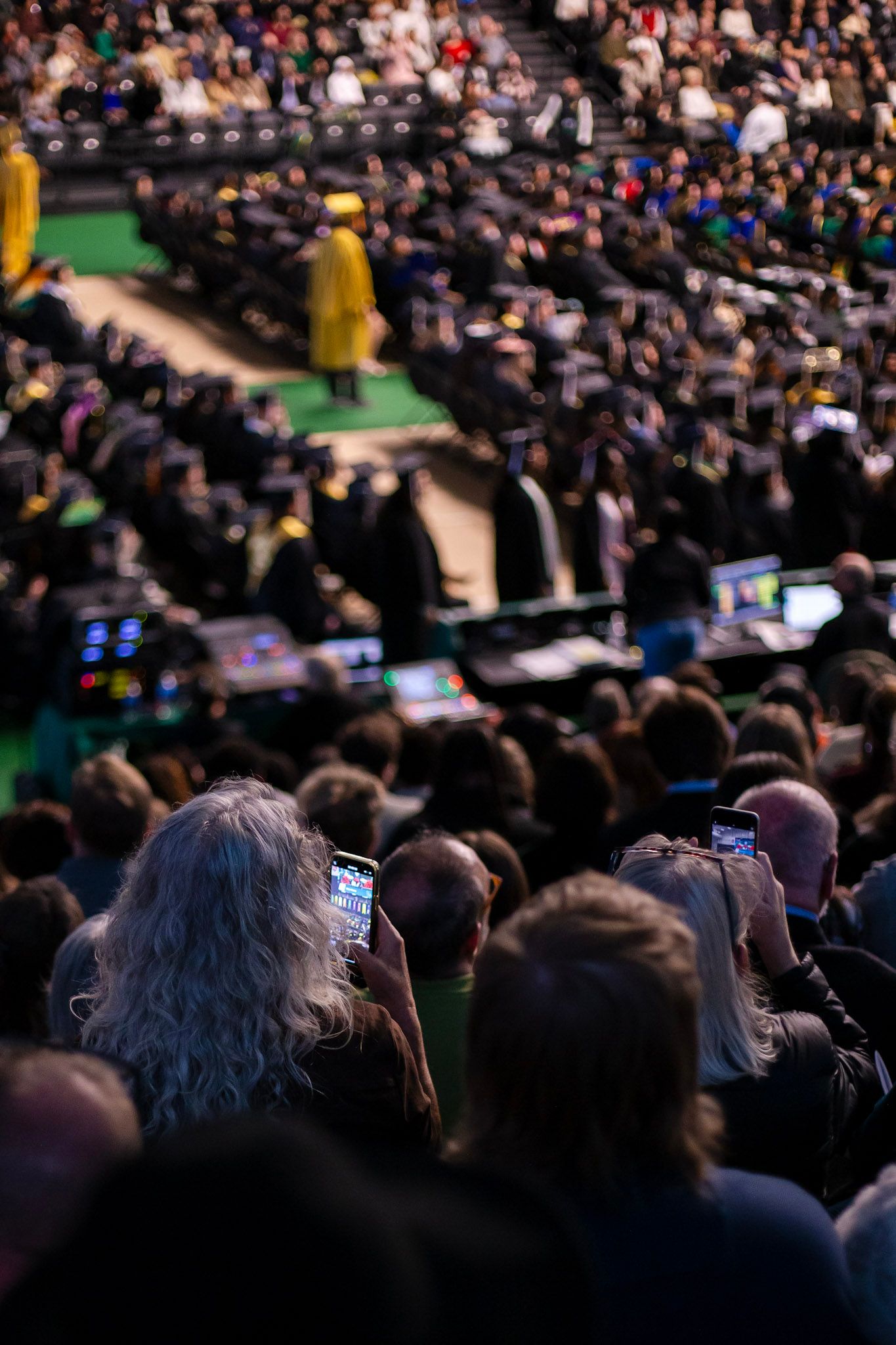 Audience members photograph graduation ceremony on smartphones as graduates fill arena floor below.