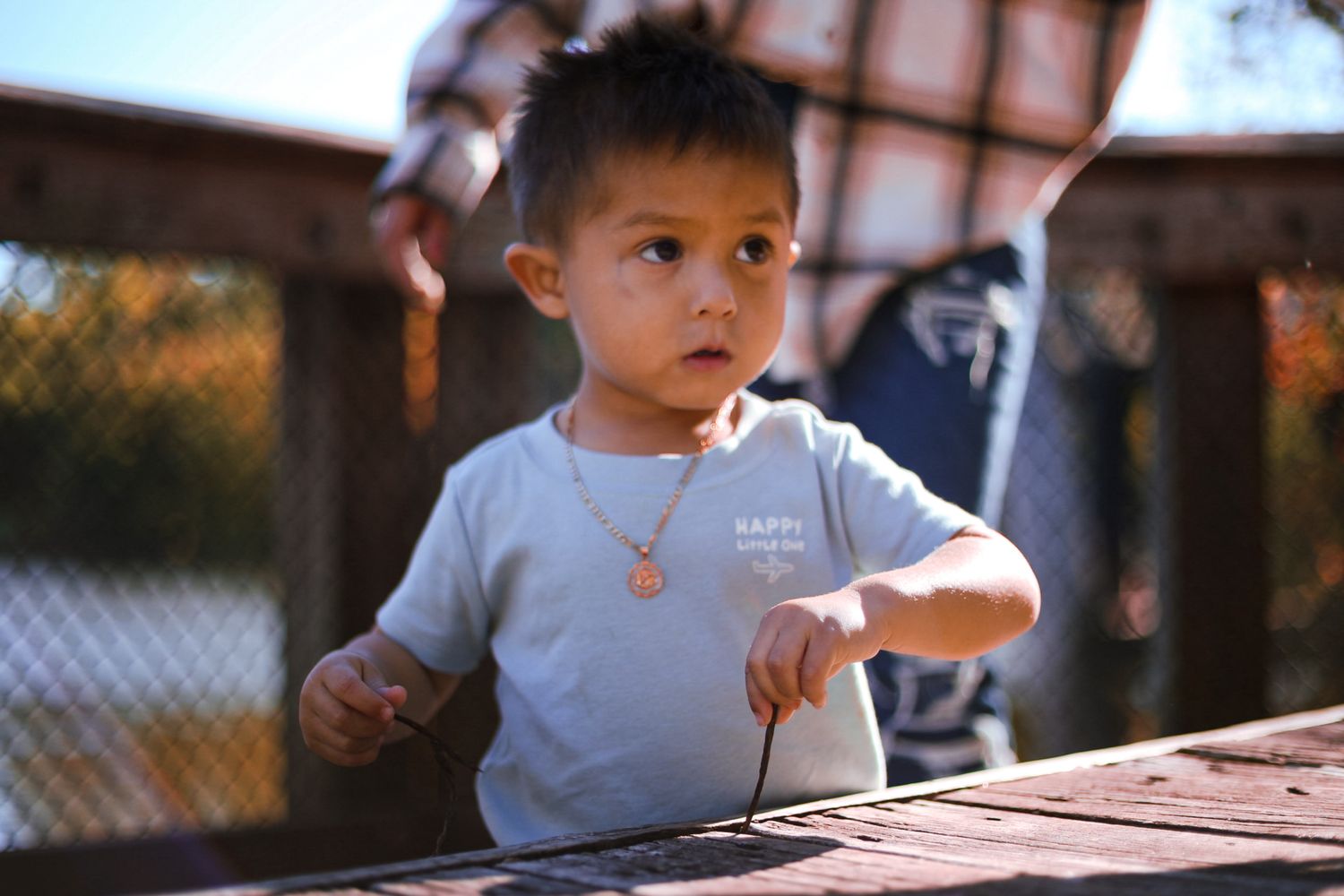 Curious toddler boy wearing a white t-shirt and necklace standing at a wooden table outdoors.