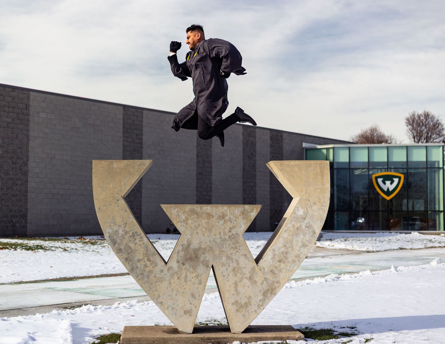 Excited Wayne State graduate jumping over large concrete W sculpture on snowy campus grounds.