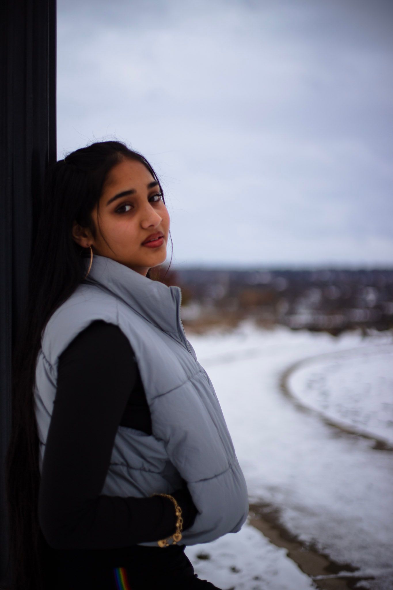 Young woman in silver puffer vest leans against dark wall with snowy landscape behind her.