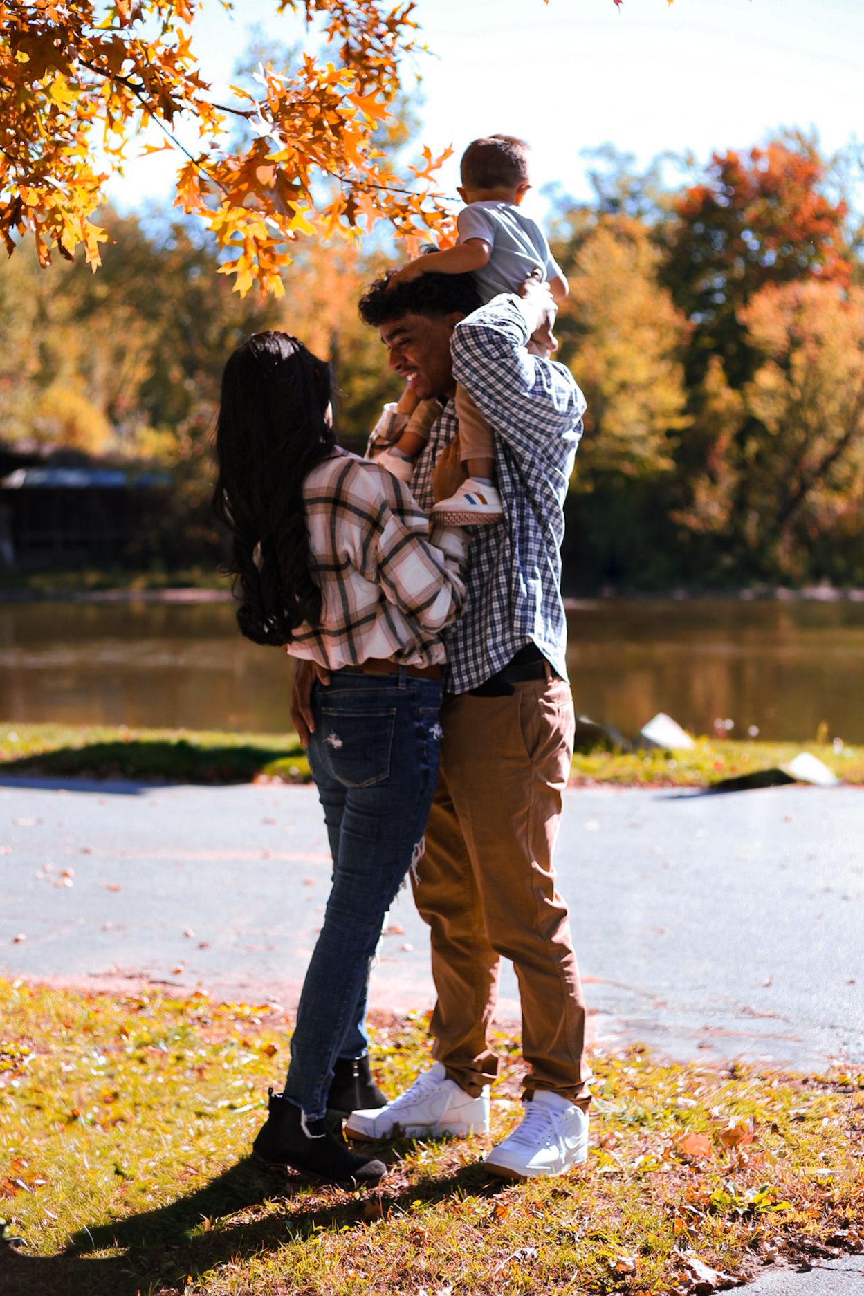 A young family with a toddler on dad's shoulders stands together by a lake in autumn scenery.