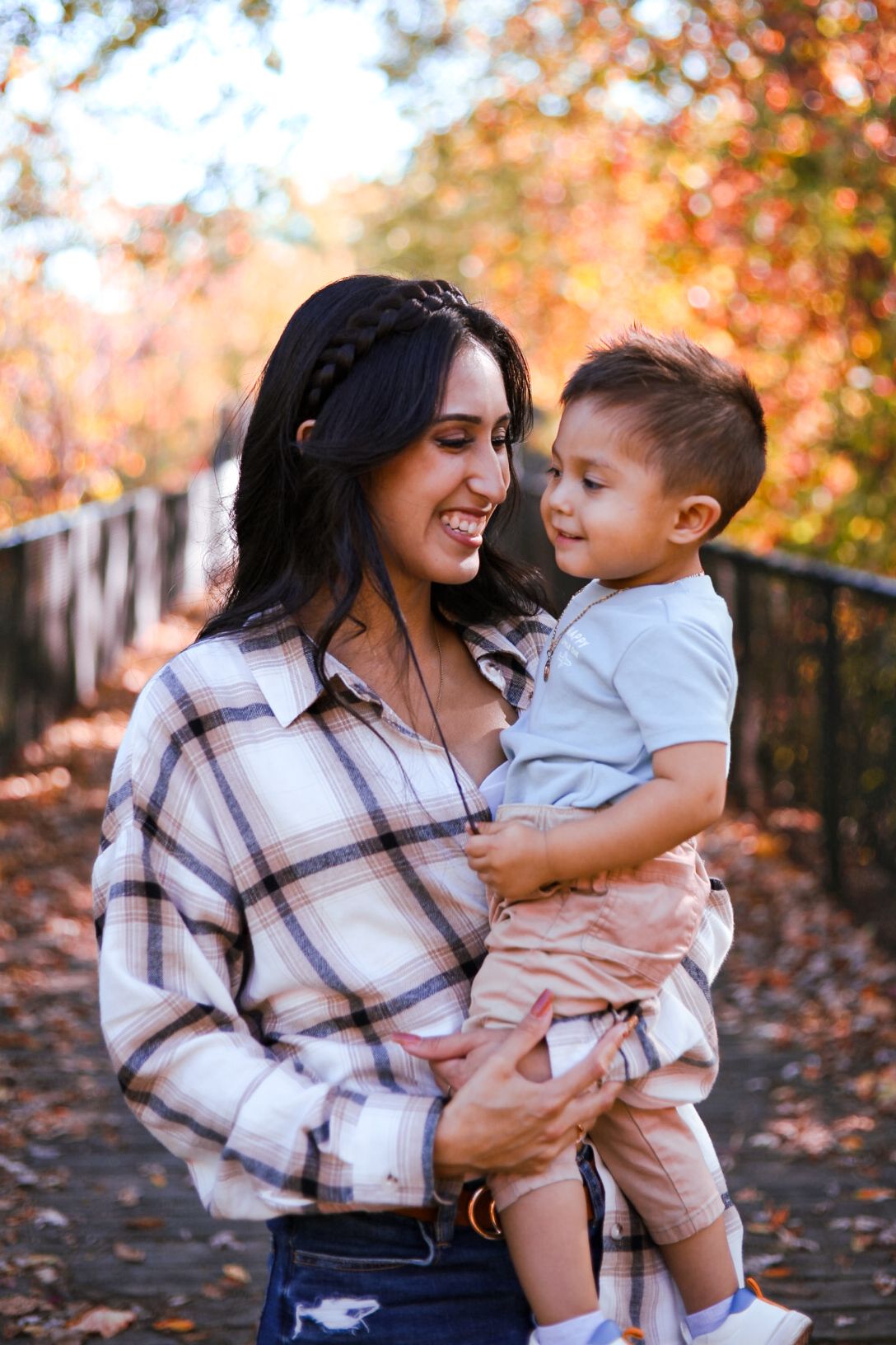 A smiling mother holds her toddler son on a bridge surrounded by colorful fall foliage.