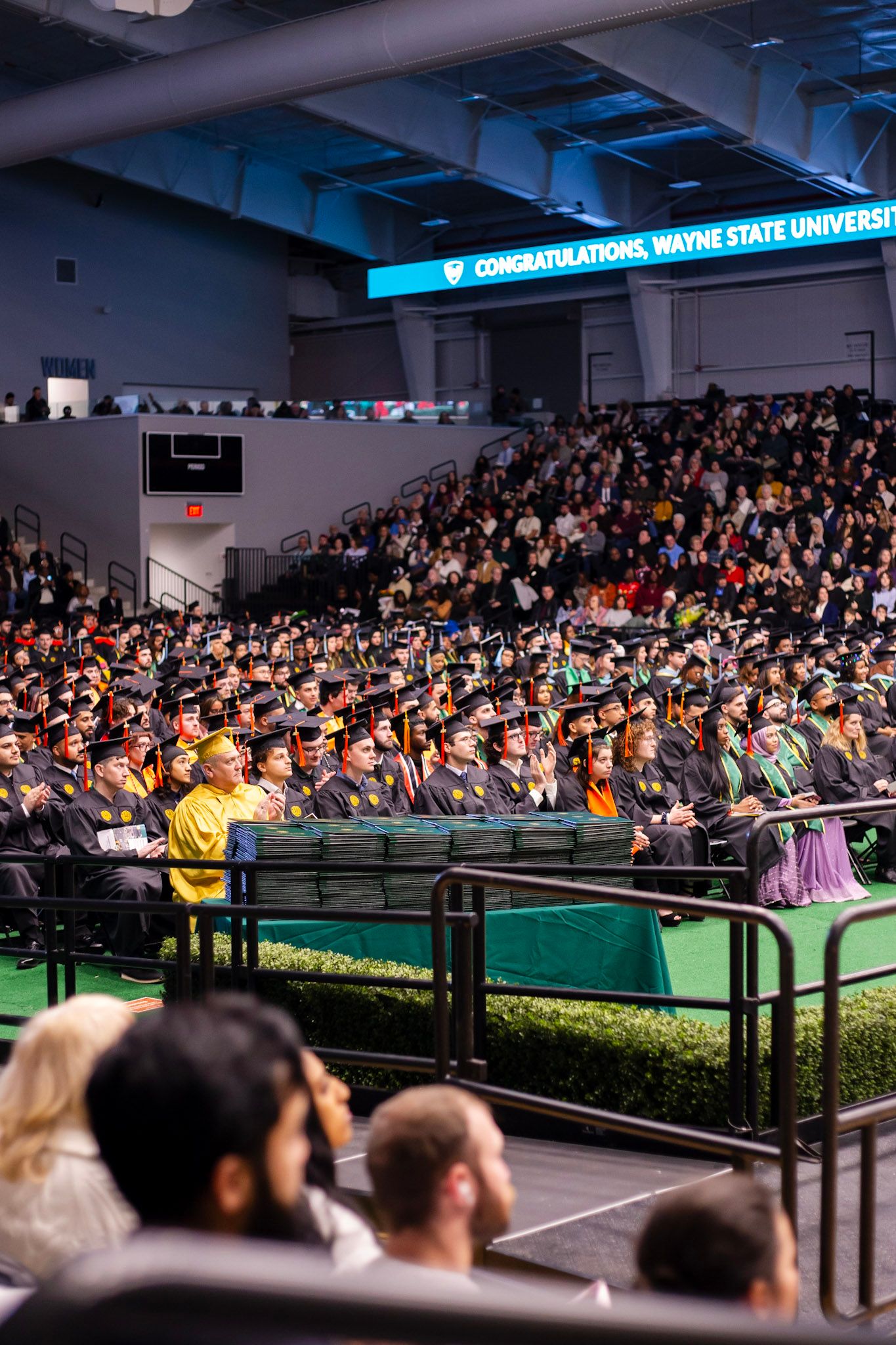 Large group of Wayne State University graduates in black caps and gowns seated at an indoor commencement ceremony.