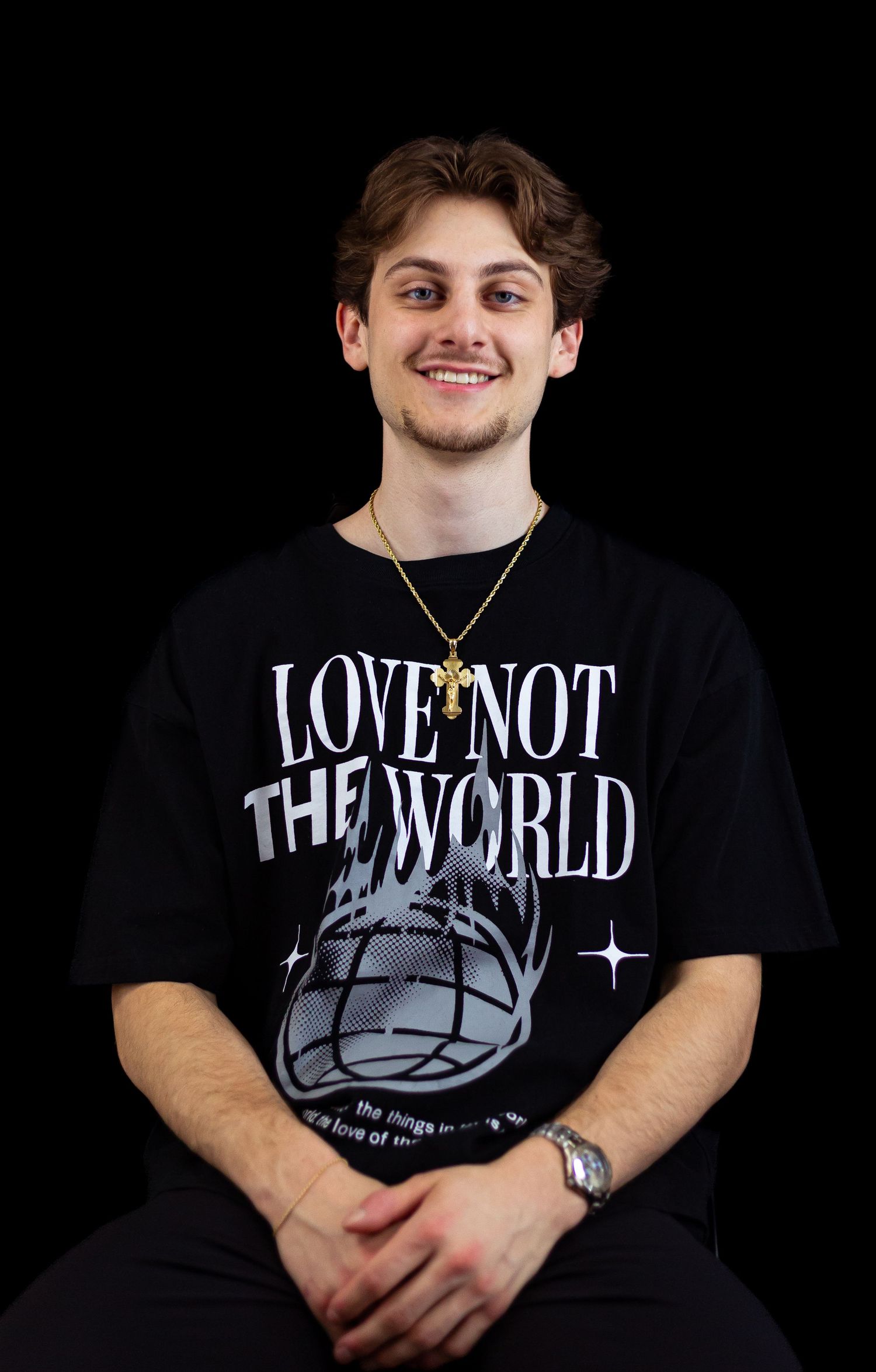 Smiling young man wearing a black Love Not The World tee and gold cross necklace sits on a black background.