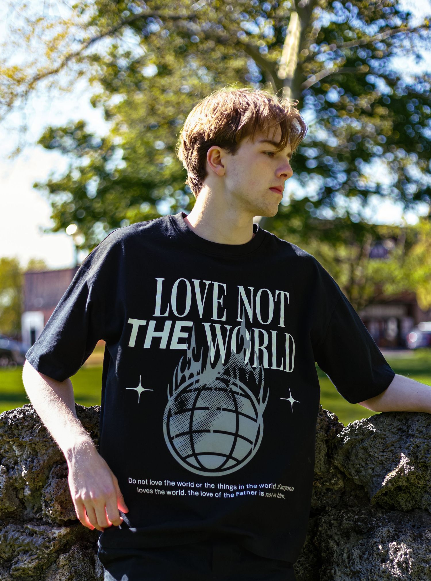 Young man leaning on stone wall wearing black Love Not The World graphic t-shirt in park.