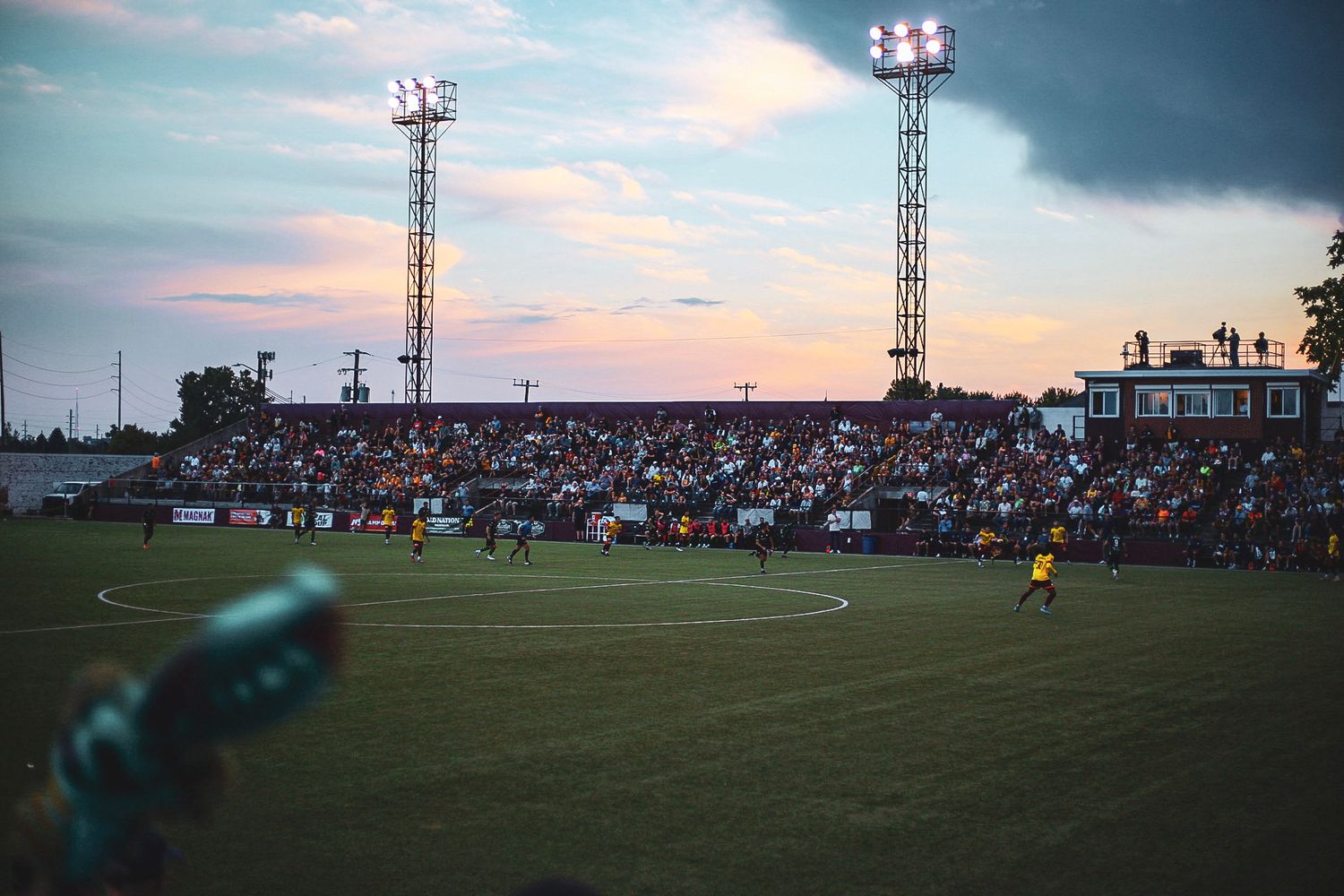 Detroit City FC soccer match at dusk with floodlit stadium packed with fans under a colorful sunset sky.