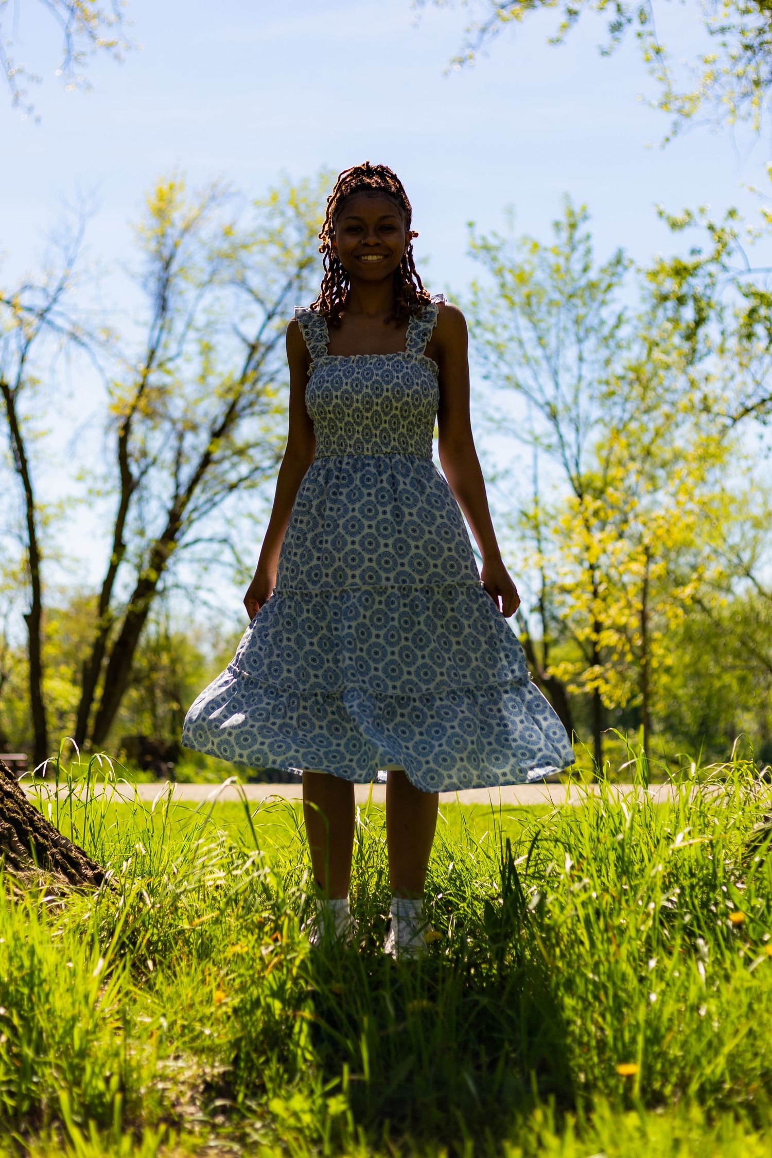 Smiling young Black woman in blue floral dress stands in a sunny green park with trees in the background.