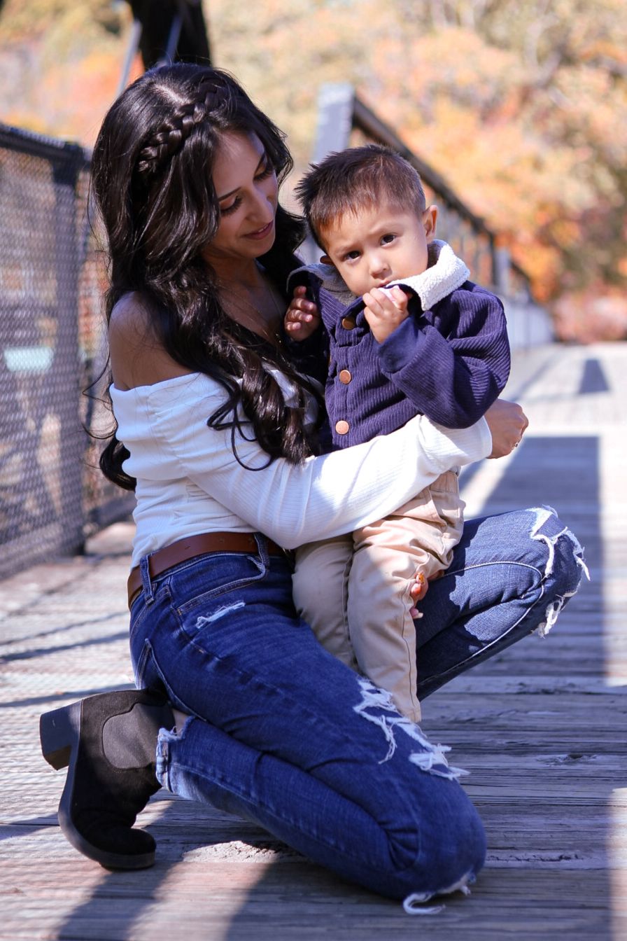 Young mother sitting on wooden bridge holding toddler son during autumn outdoor photo session.