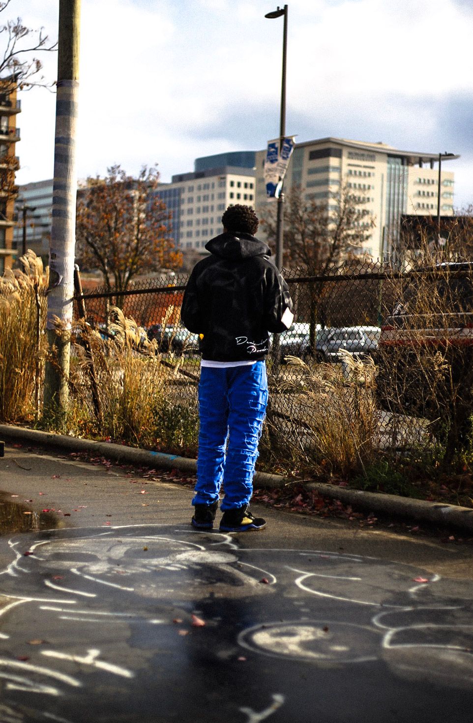 Person in blue pants and black jacket standing on urban sidewalk with city buildings in background.