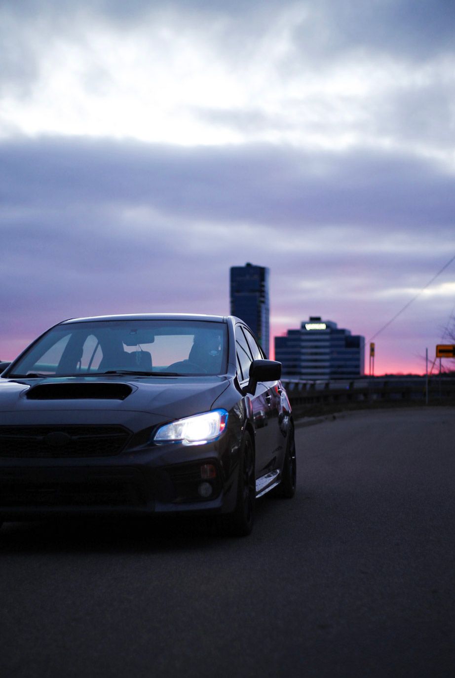 Dark Subaru WRX with headlights on parked at dusk under a dramatic purple and pink cloudy sky.