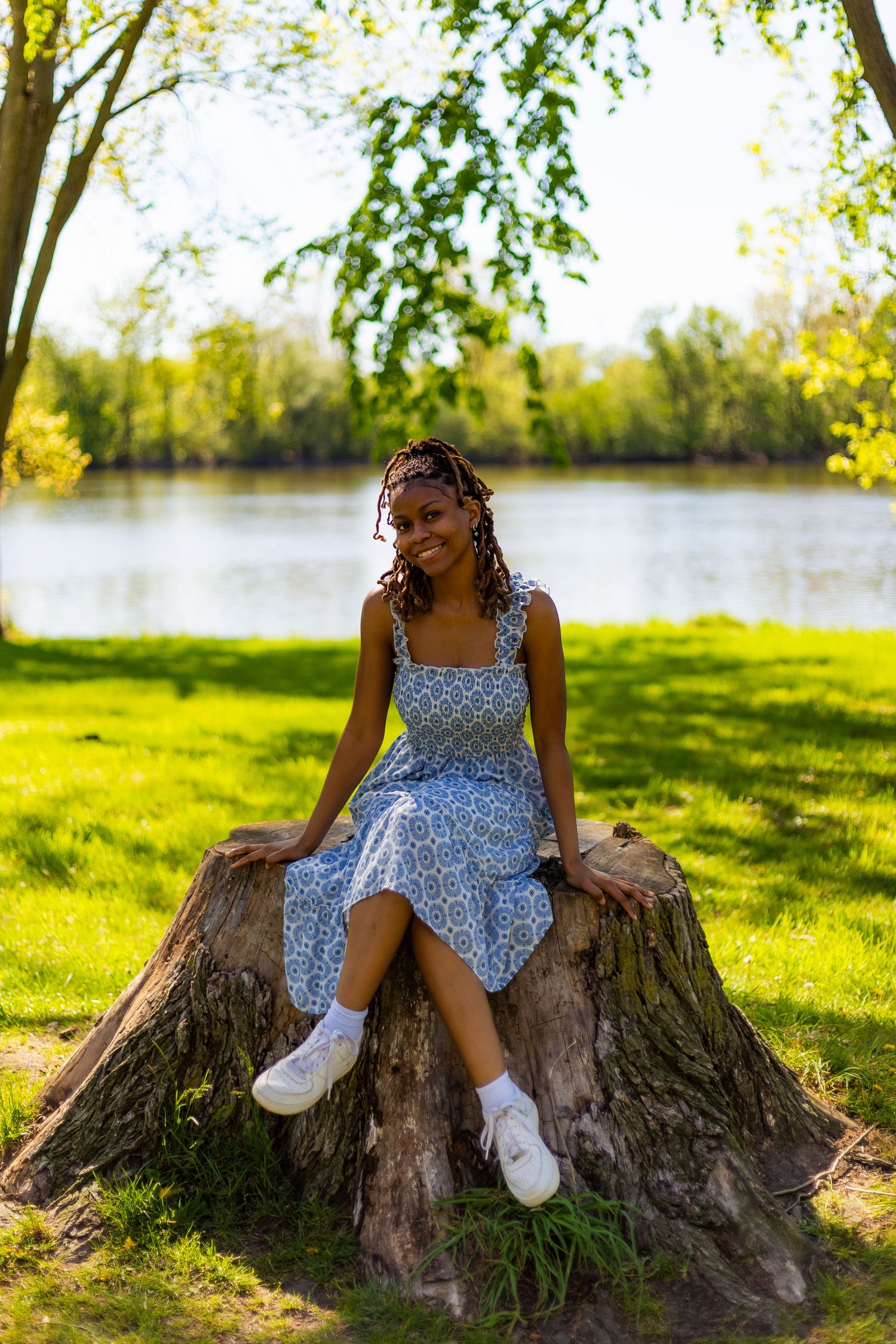 Smiling young Black woman in blue floral dress sits on large tree stump by lake.