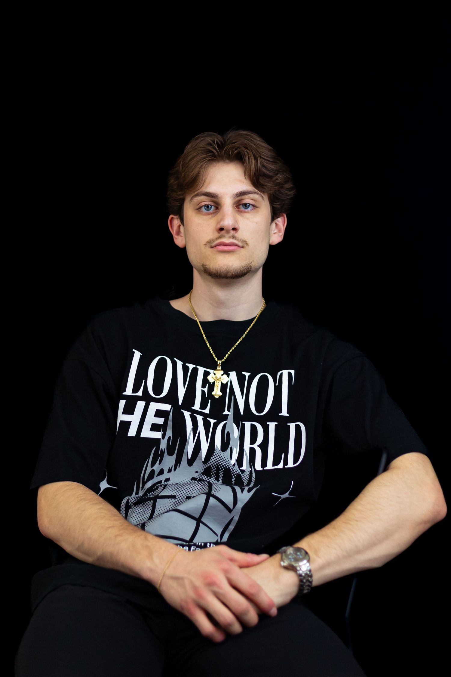 Young man in black Love Not The World t-shirt with gold cross necklace seated against black background.