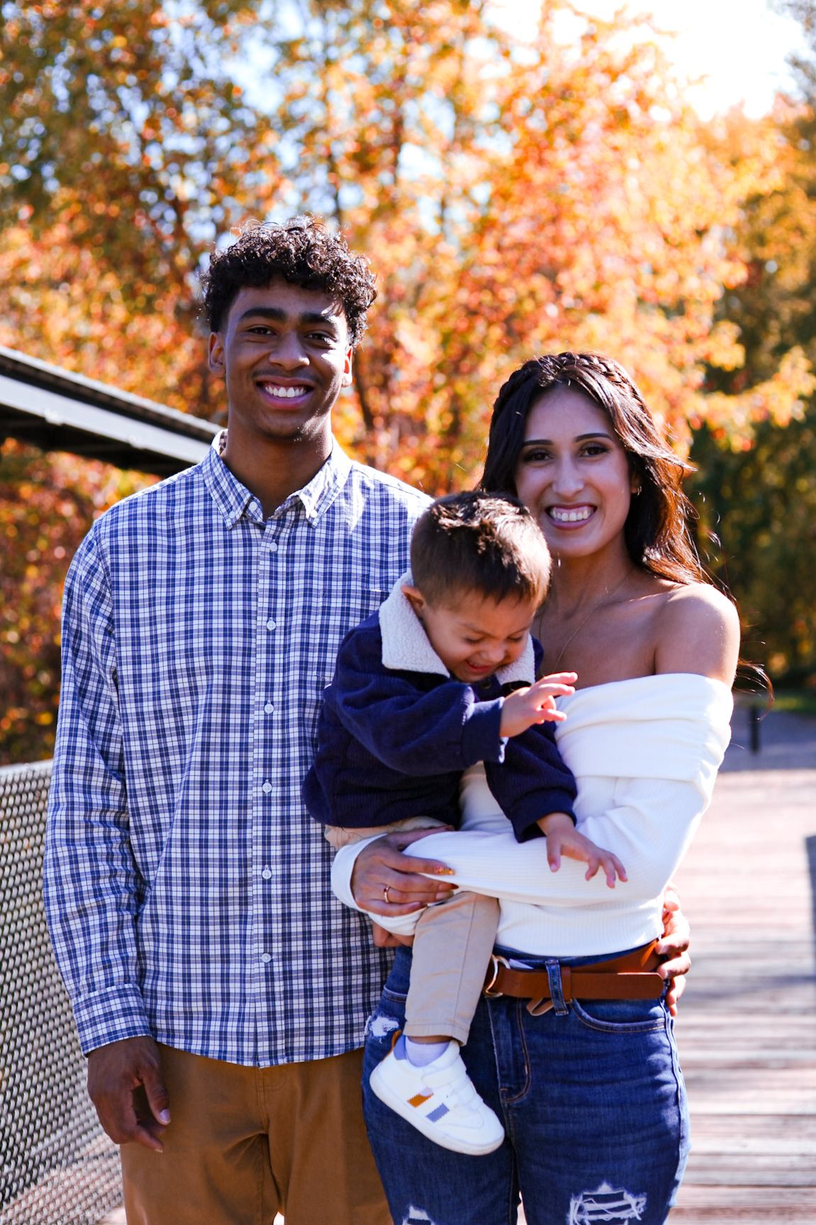 Smiling young family posing together on bridge with colorful autumn foliage in background.
