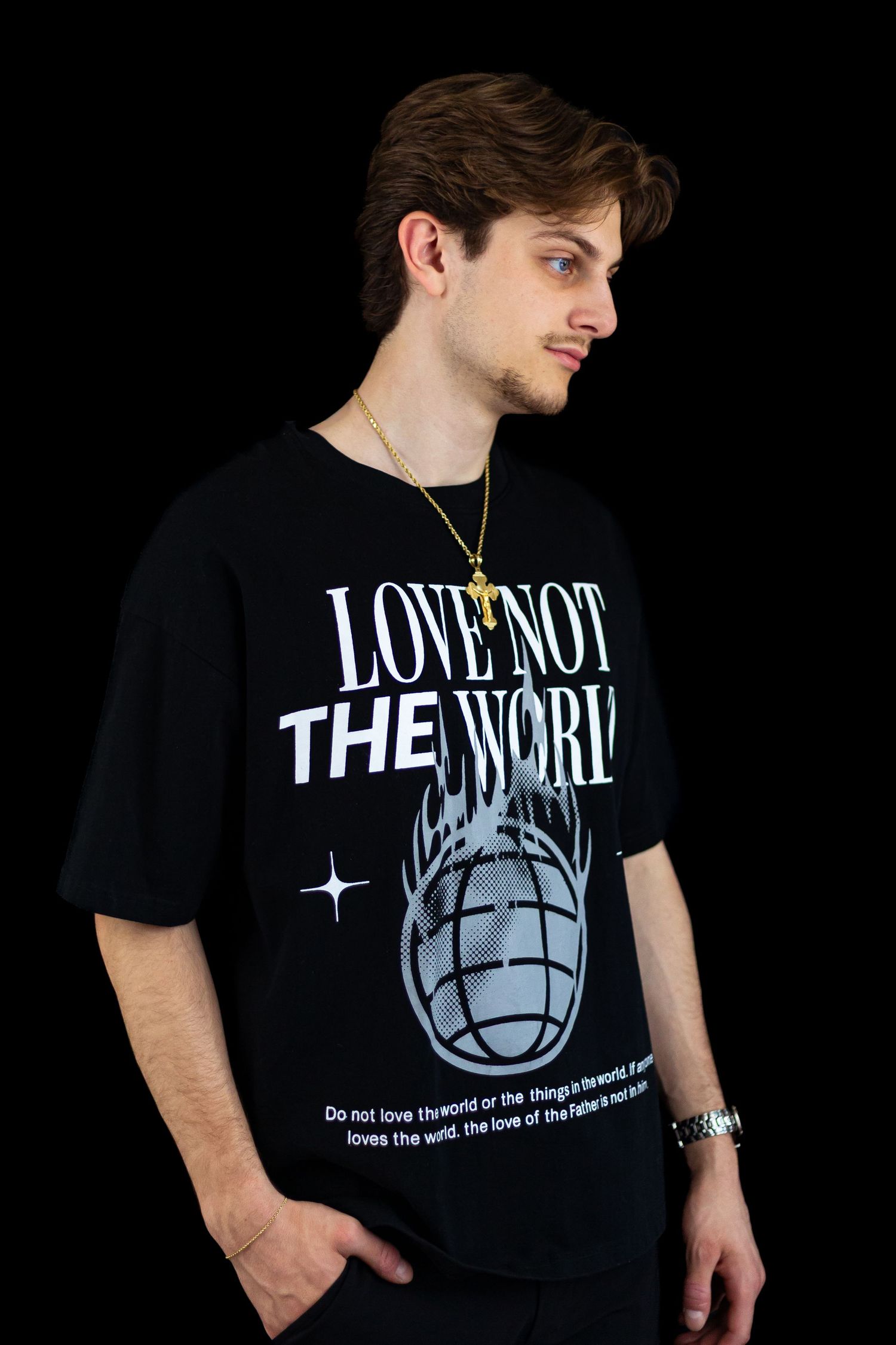 Side profile of young man in black Love Not The World graphic tee with gold cross chain necklace.