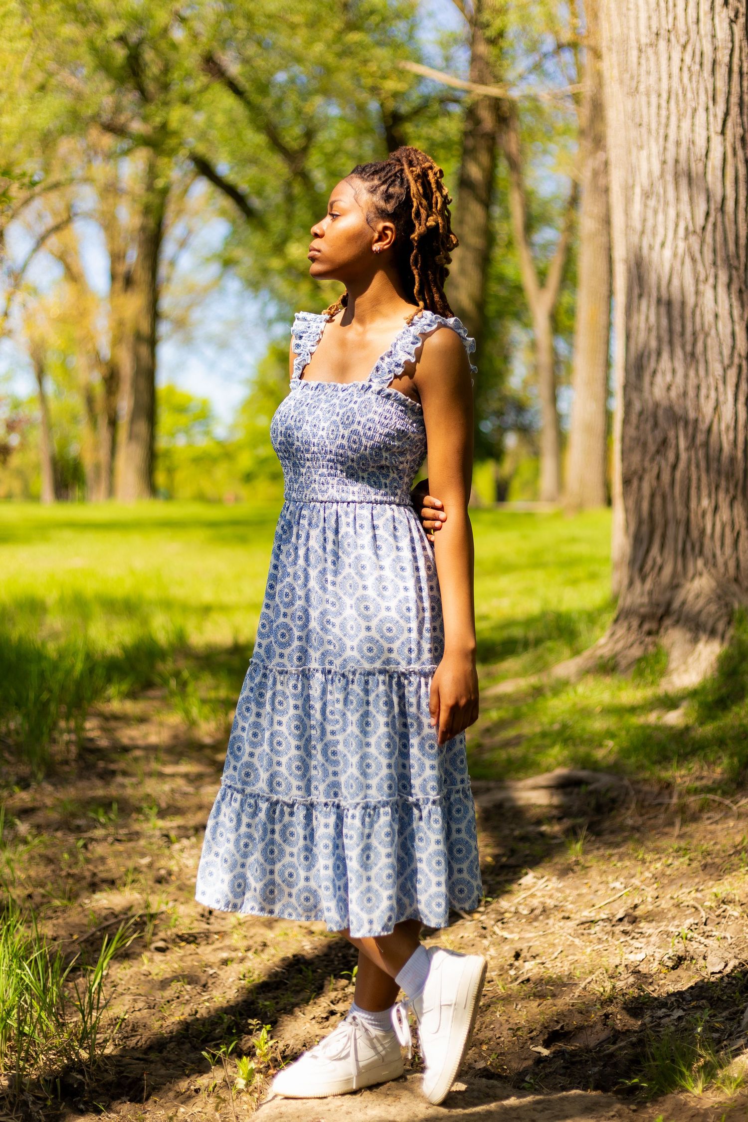 Young Black woman in blue patterned midi dress stands on a sunlit park path surrounded by tall trees.
