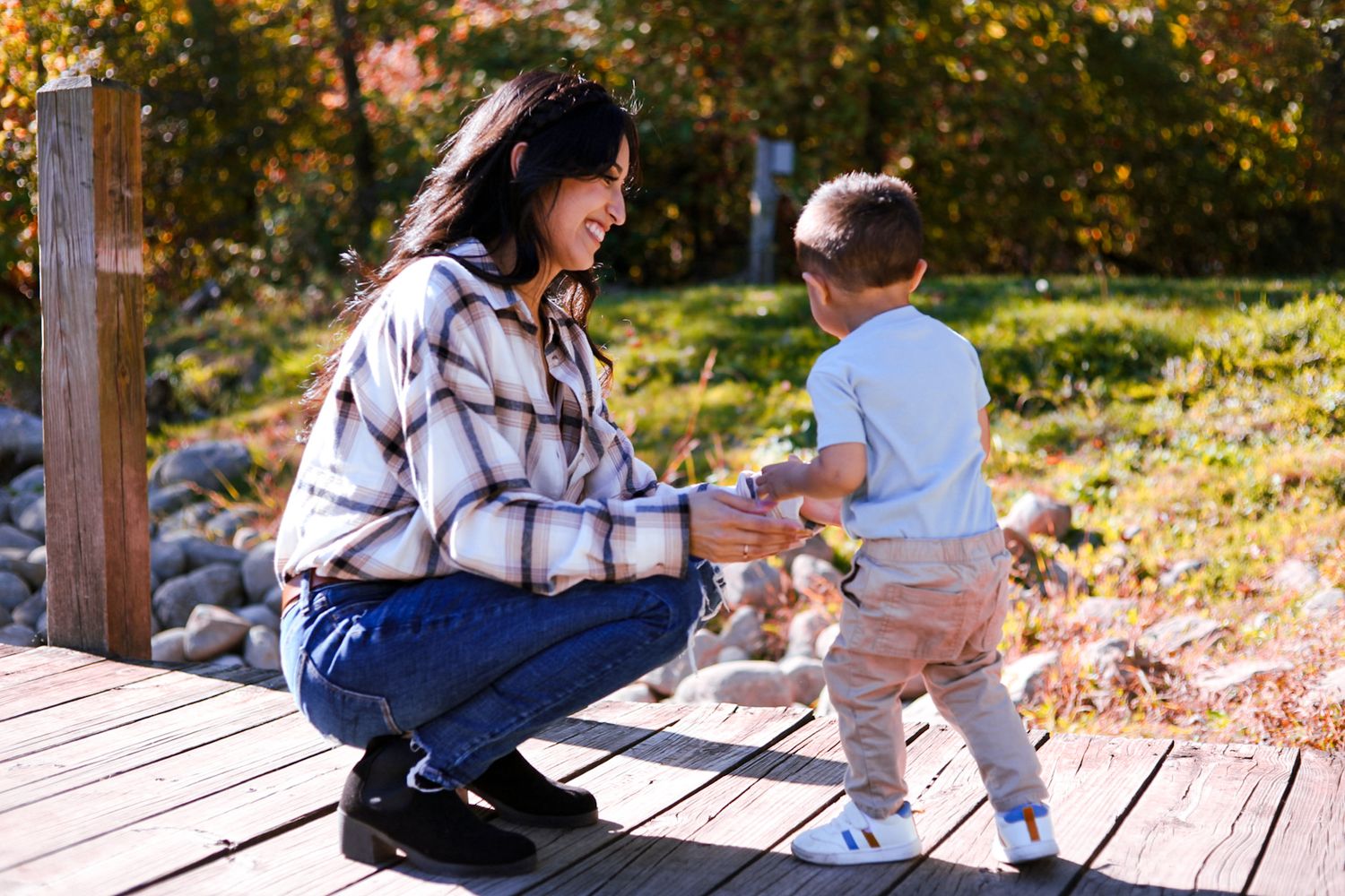 A smiling mother kneels on a sunny wooden dock holding her toddler son's hands in autumn.