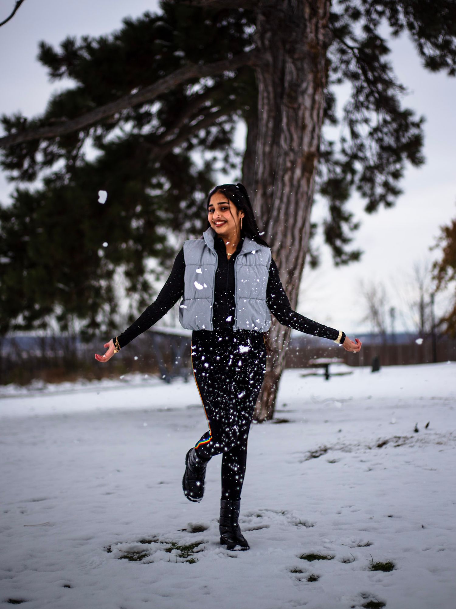 Joyful young woman in silver vest plays in falling snow near trees in winter park.