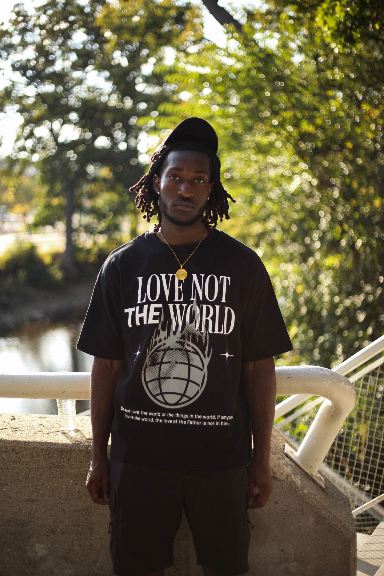 Man in black 'Love Not The World' tee wearing black cap and gold chain posing on sunny bridge.