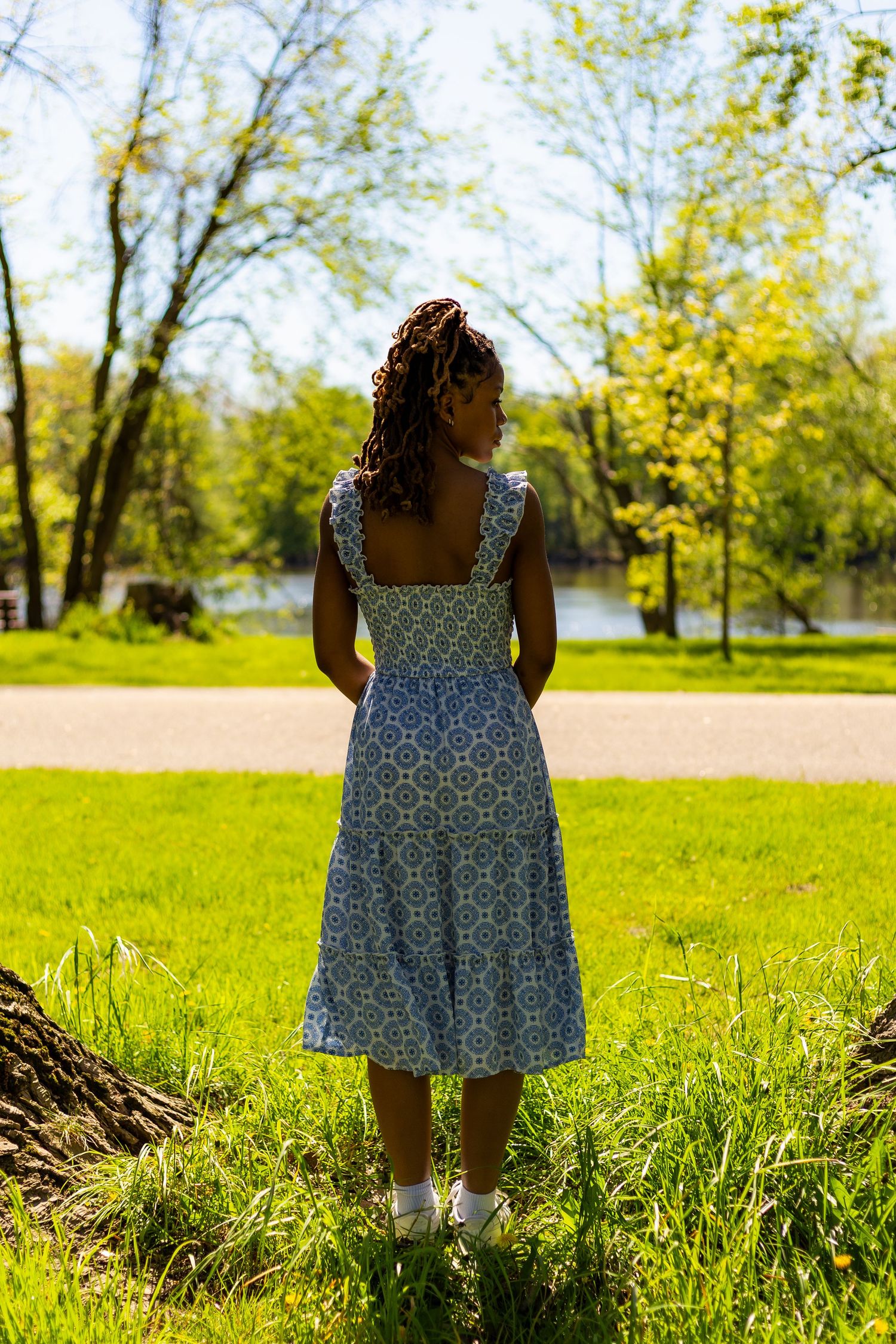 Rear view of young Black woman in blue floral sundress standing in a green park looking toward a lake.