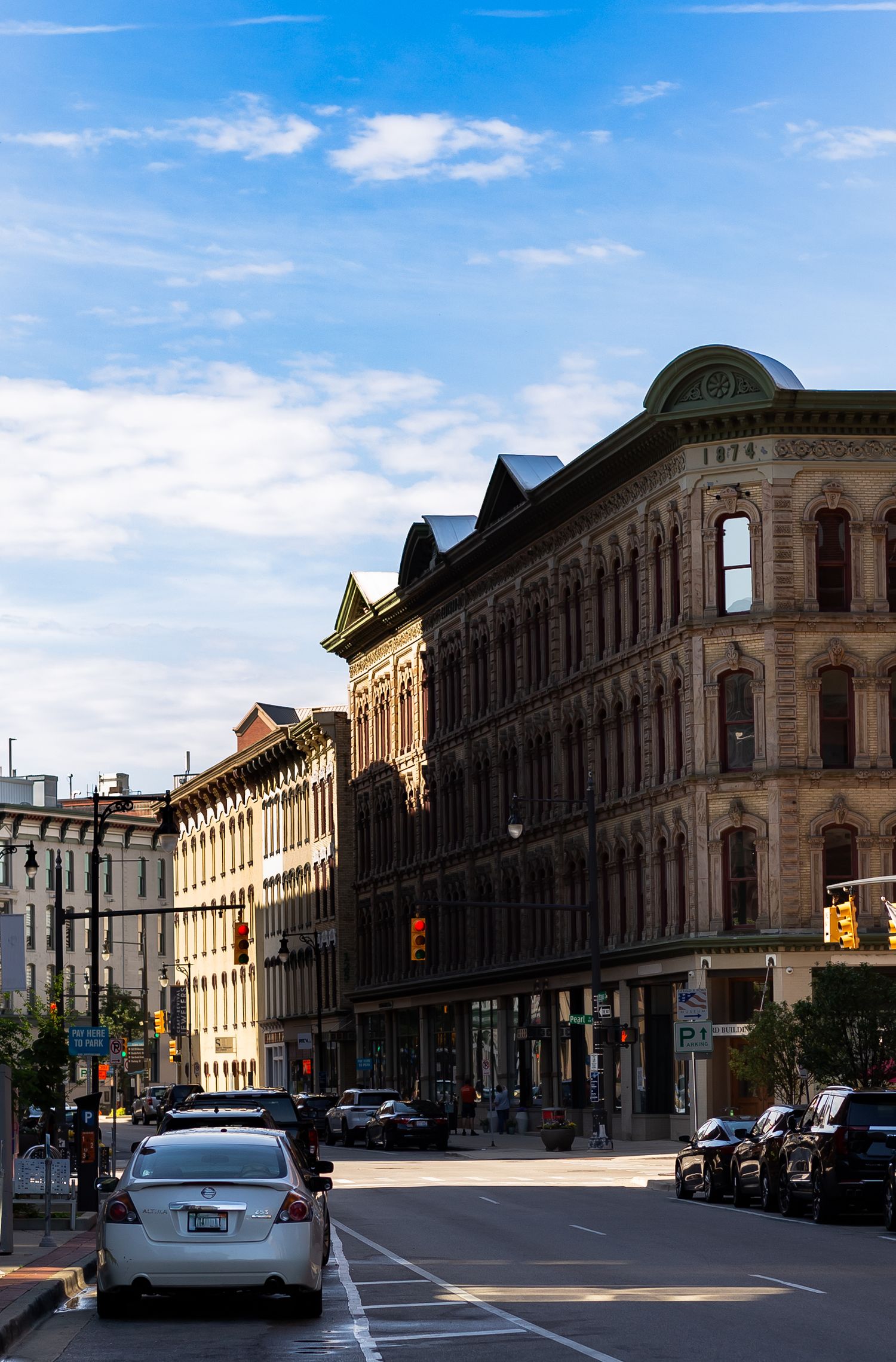 Historic brick buildings line a sunny urban street with parked cars and a blue sky in what appears to be a classic American city downtown.