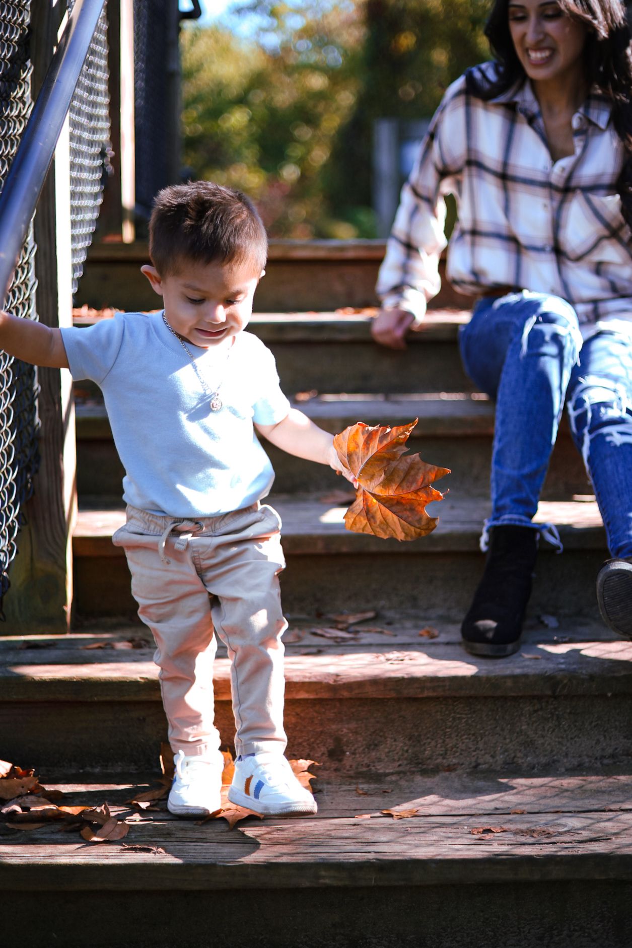 Toddler boy holding autumn leaf on wooden steps while mother watches in fall park setting.