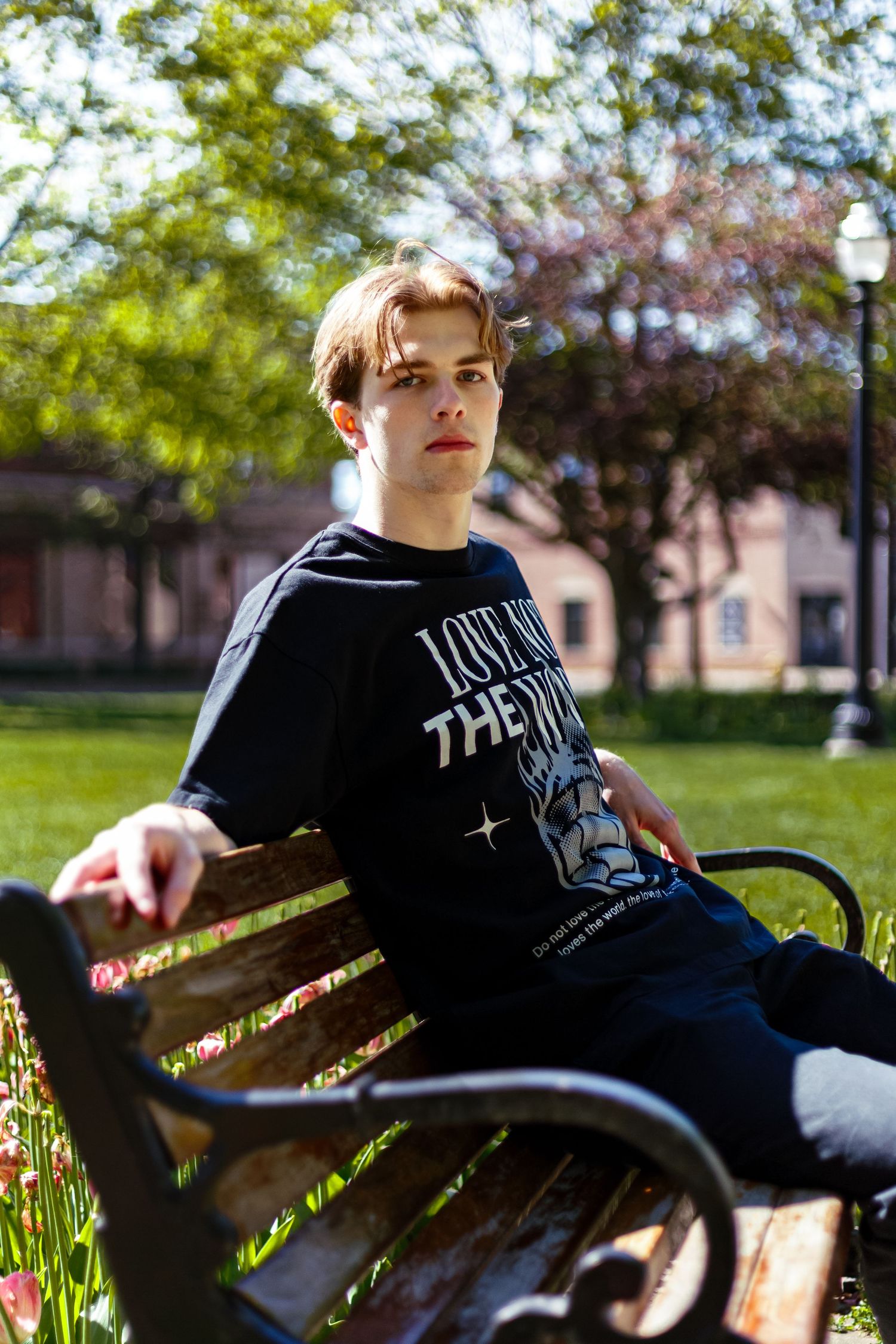 Young man sitting on wooden park bench wearing black Love Not The World graphic t-shirt.