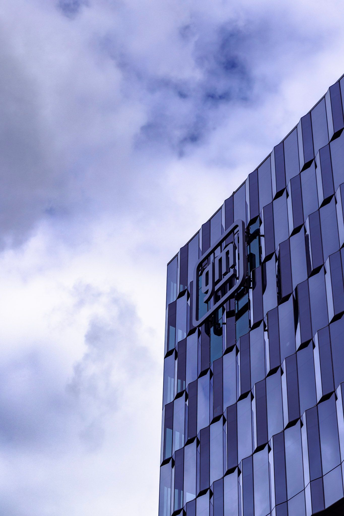 Modern glass office building with company logo reflecting cloudy blue sky from low angle view.