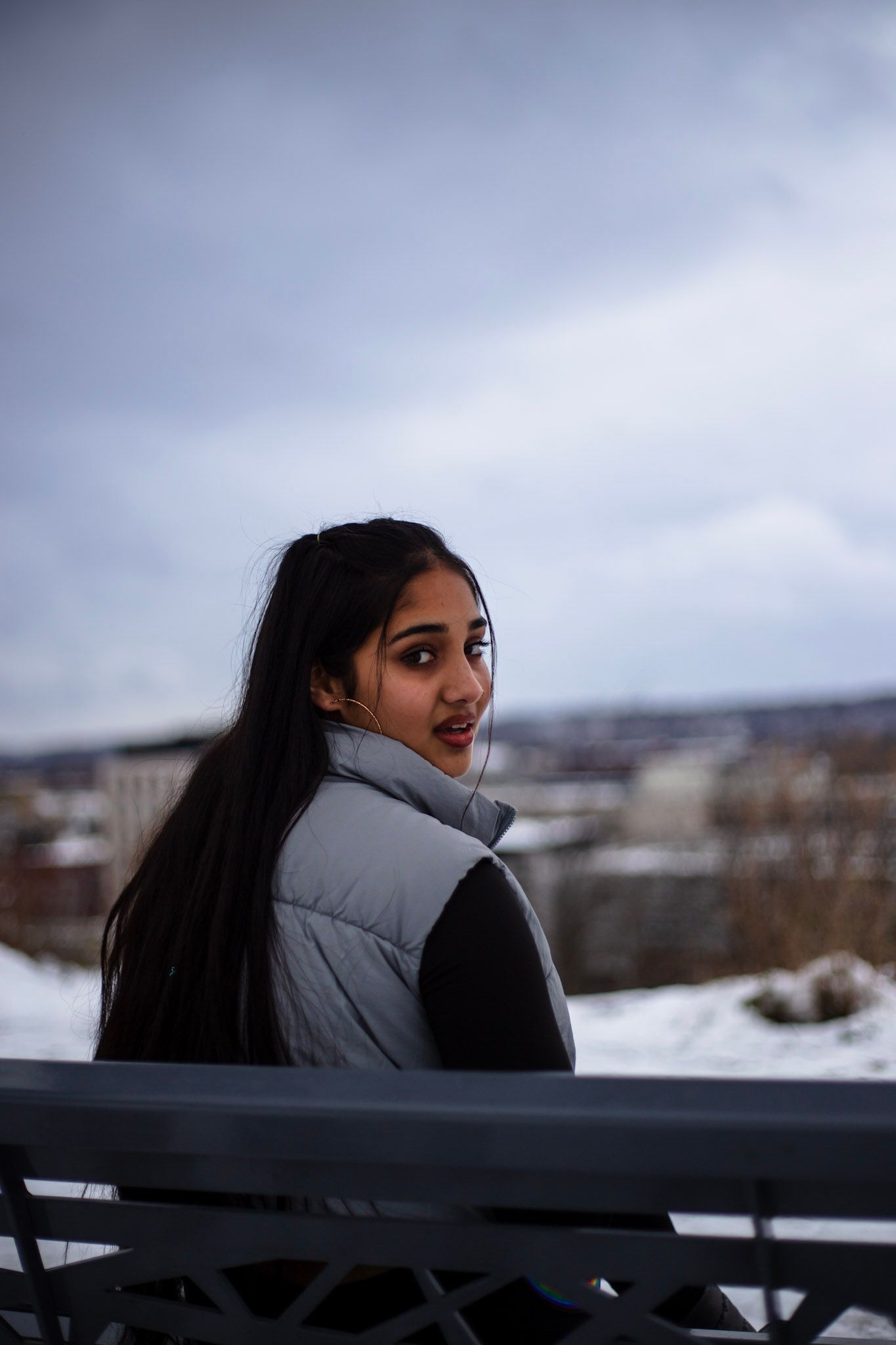Young woman in silver puffer vest sits on bench, glancing back at snowy cityscape view.