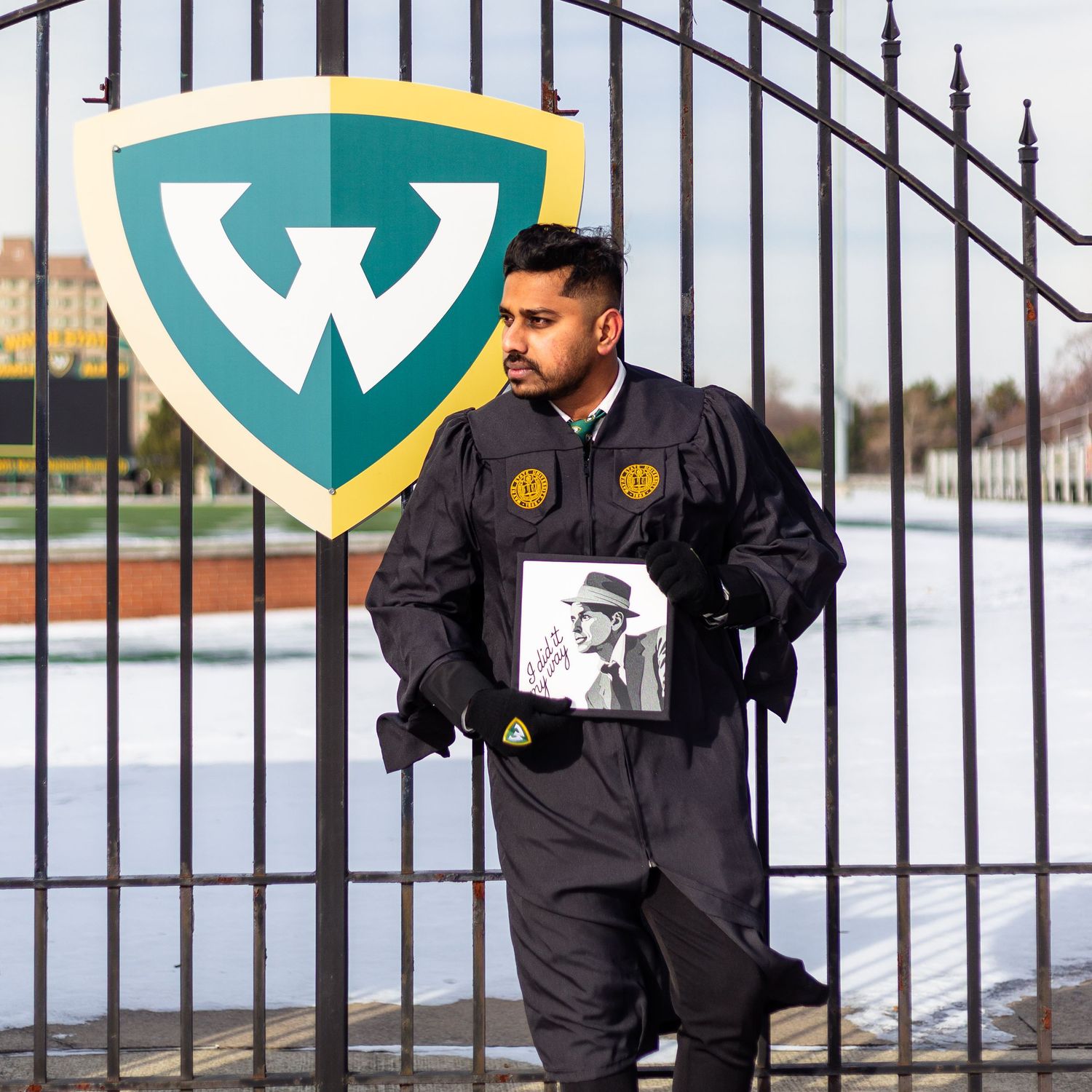 Wayne State graduate in black gown holds Frank Sinatra portrait at campus gate.