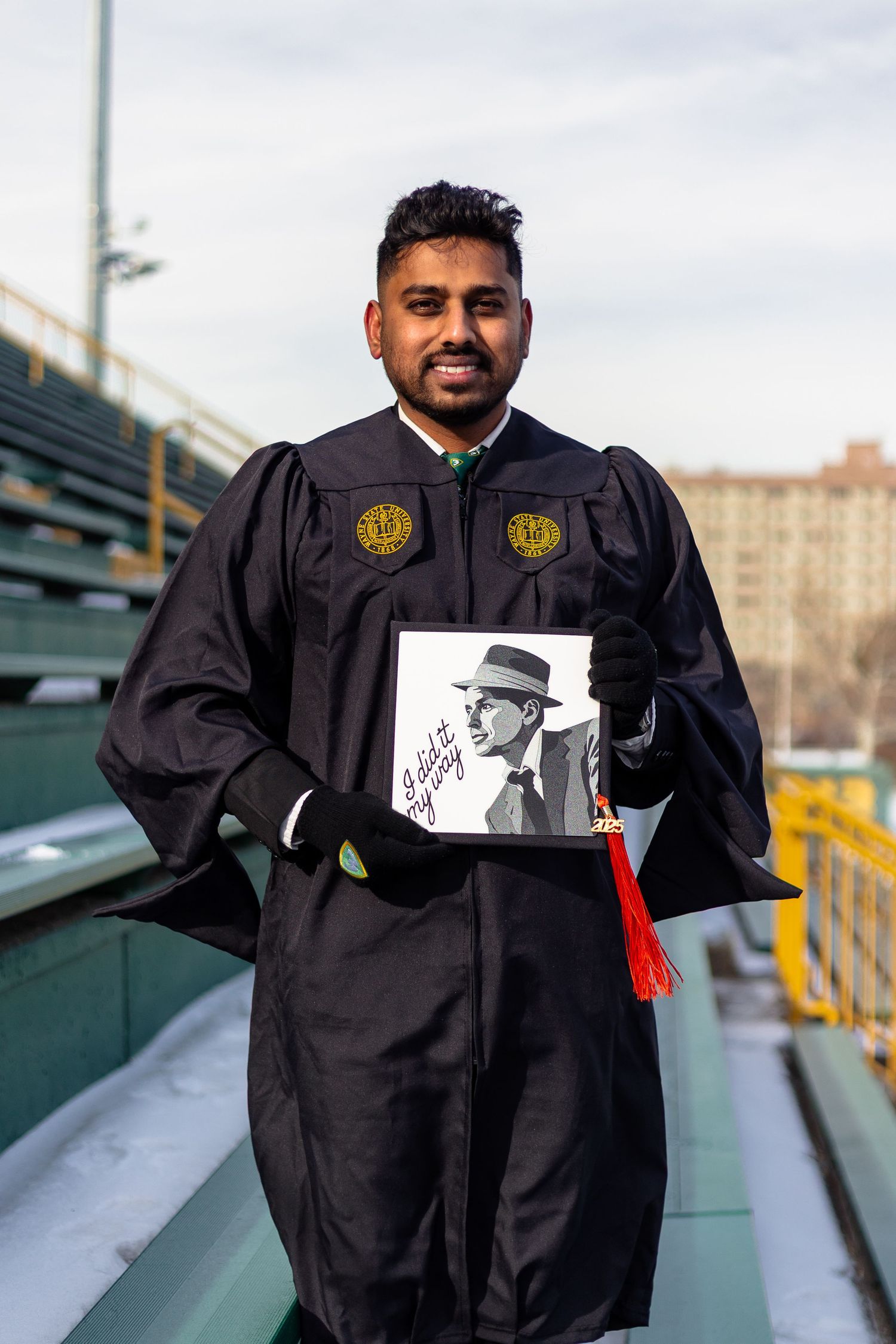 Smiling Wayne State University graduate holds Frank Sinatra decorated cap in black graduation gown.
