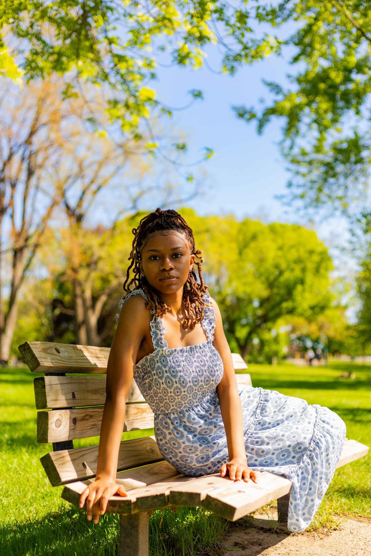 Young Black woman in a blue patterned sundress sitting on a wooden park bench surrounded by green trees.