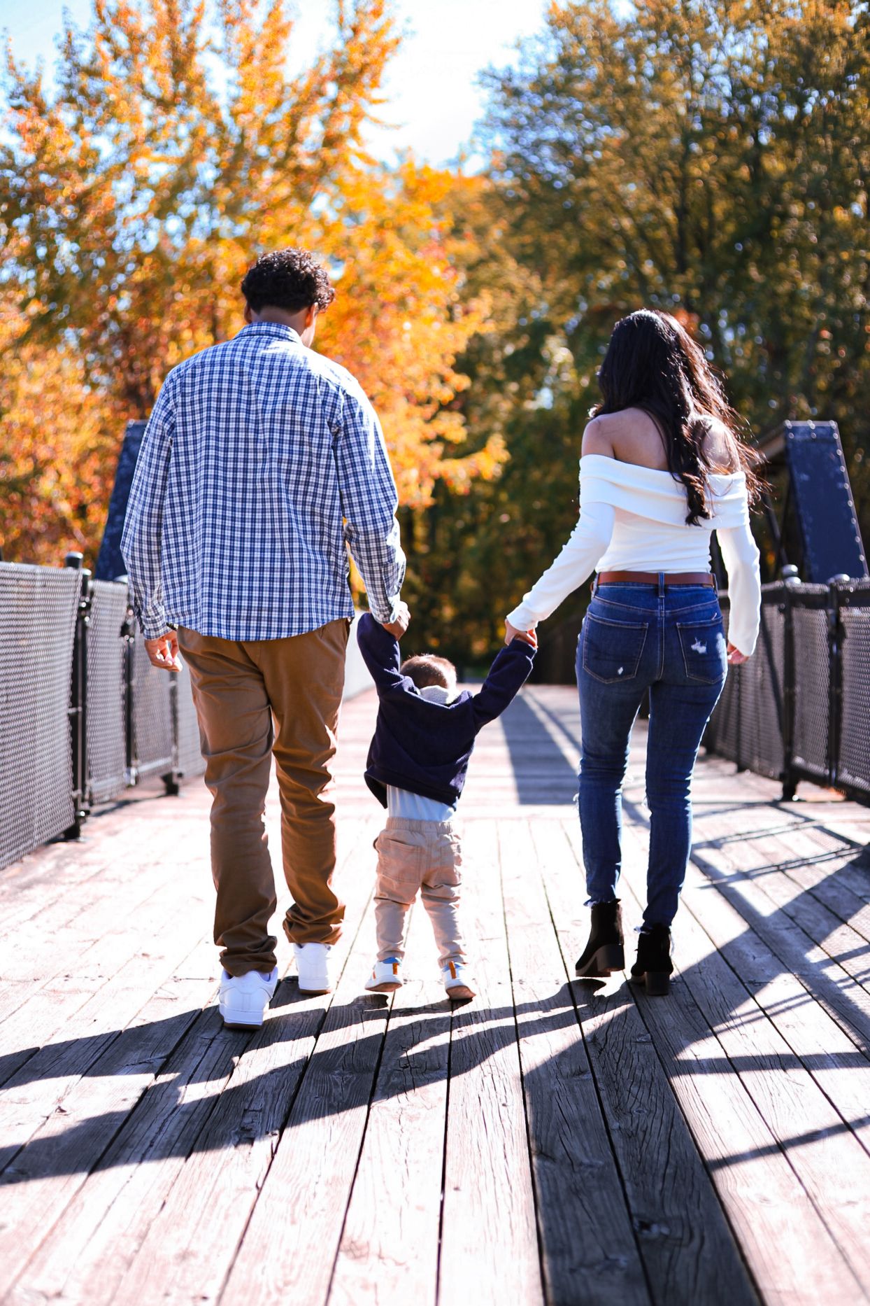 A family of three walks away hand-in-hand on a wooden bridge surrounded by colorful fall trees.