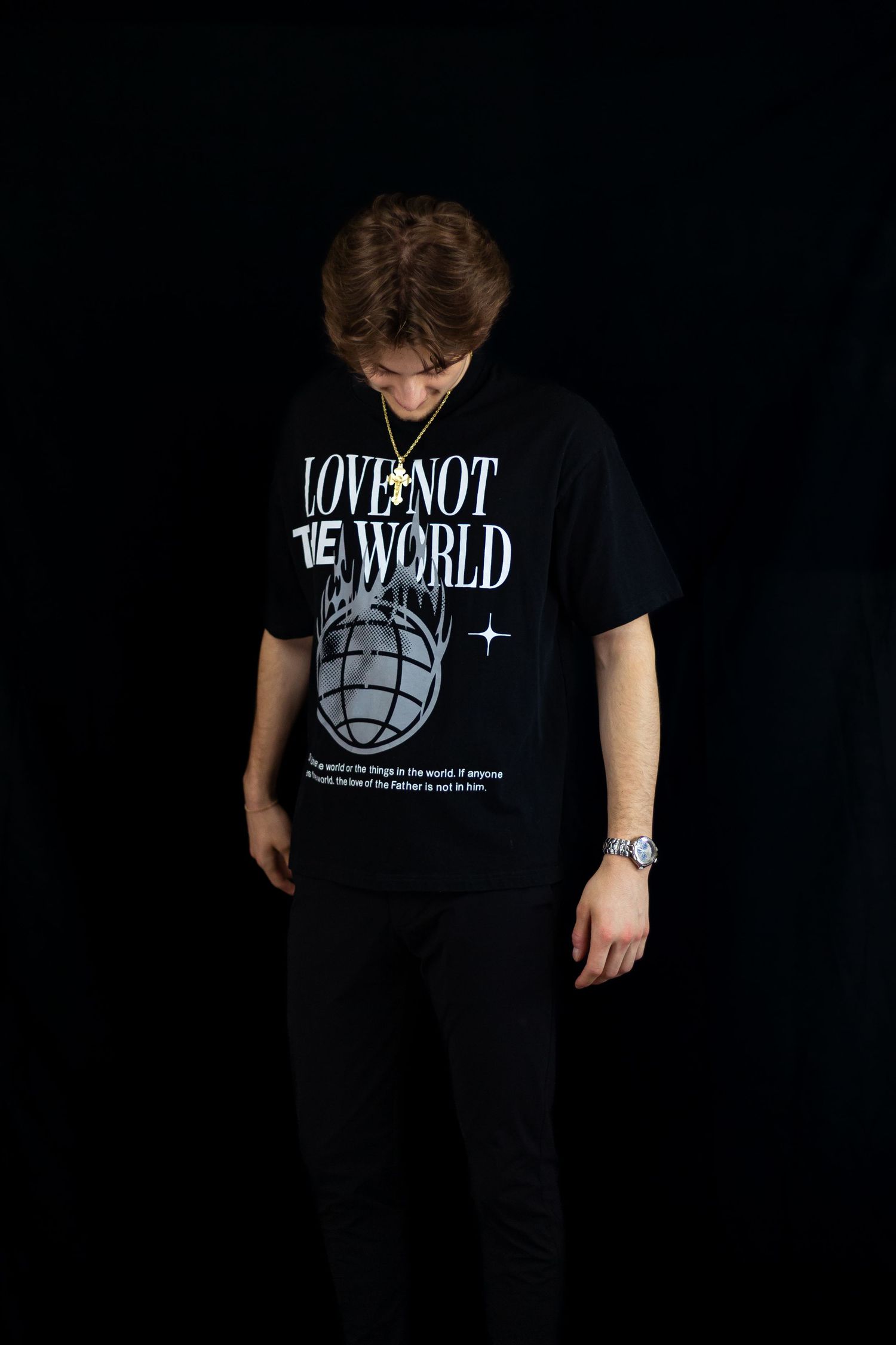 Young man looking down wearing black Love Not The World graphic tee with gold cross necklace.