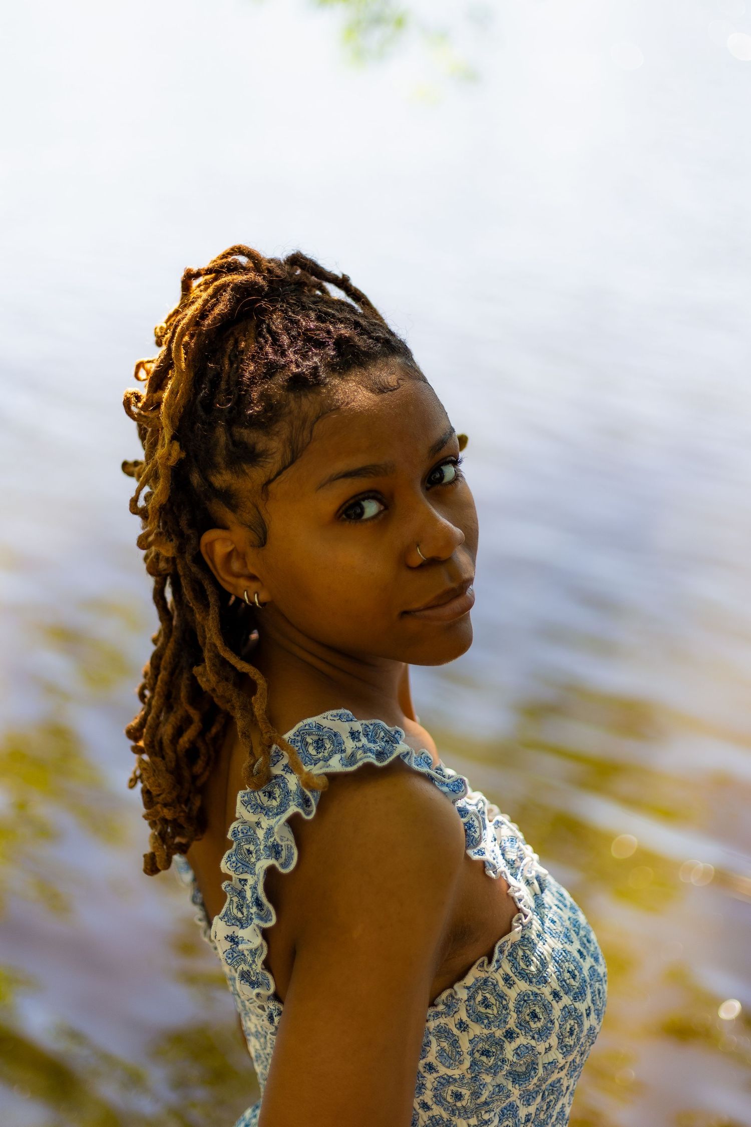 Portrait of a young Black woman with auburn locs wearing a blue floral dress near a shimmering lake.