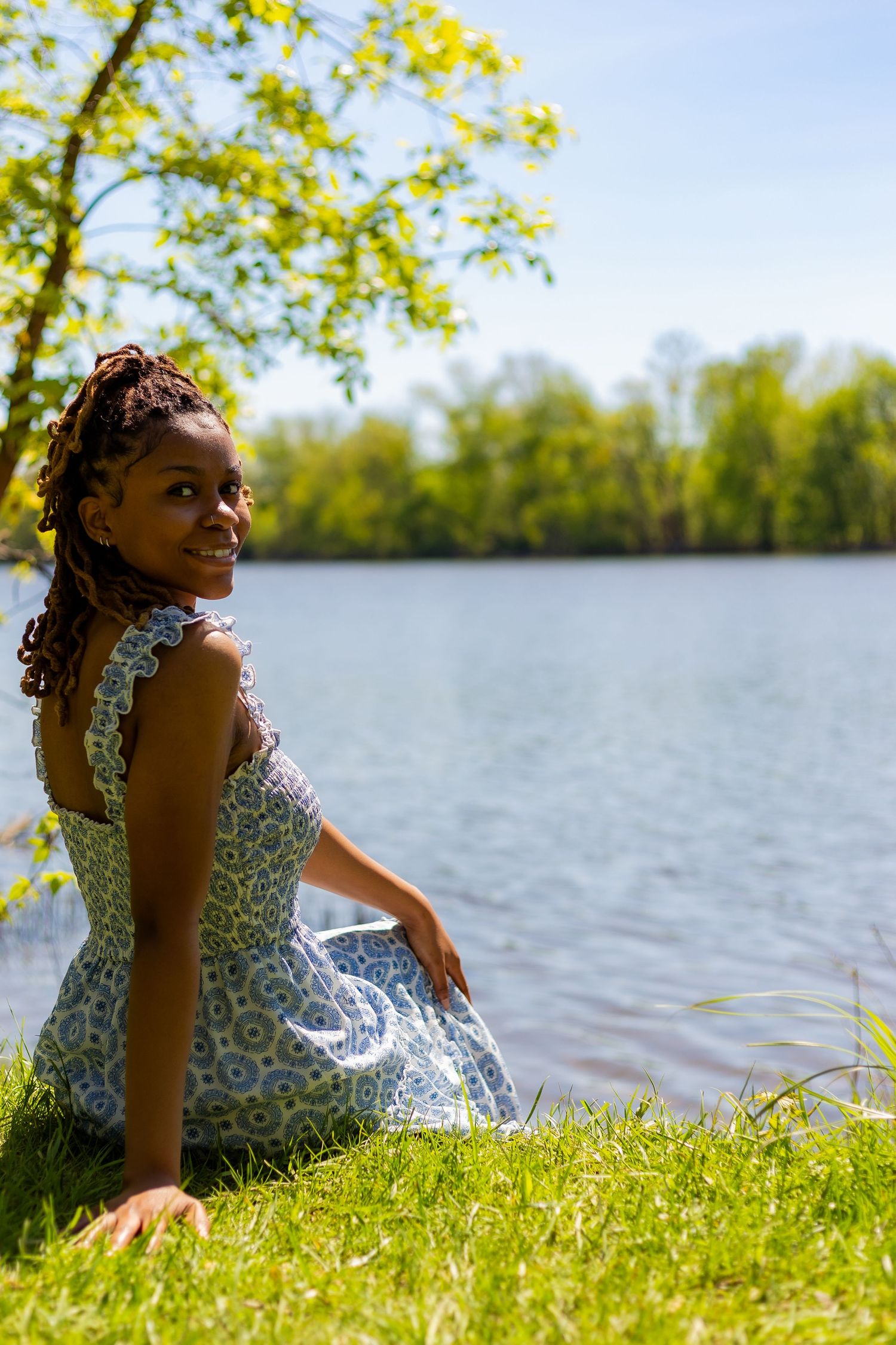 Young Black woman in floral sundress sitting on grass smiling by sunny lakeside.