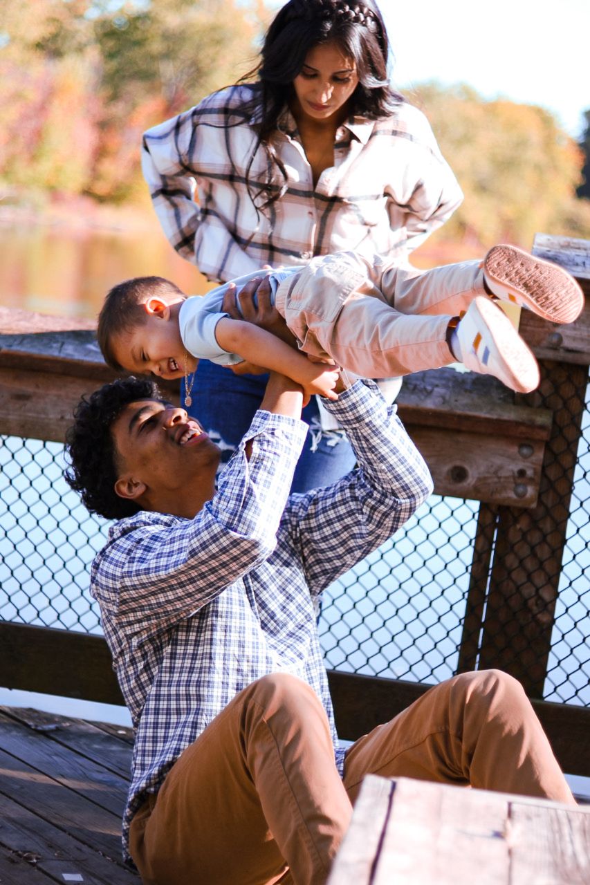 Happy family playing on wooden bridge deck with father lifting toddler while mother watches.
