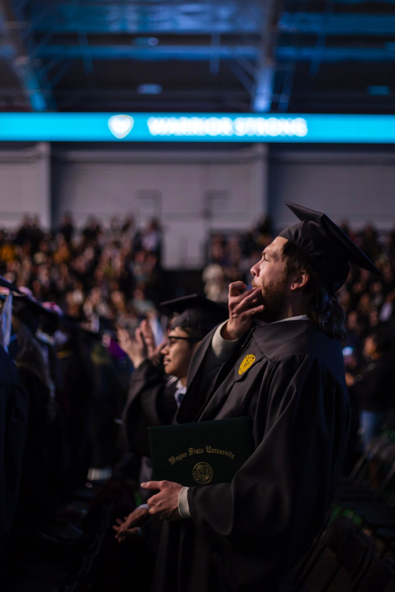 A graduate in black cap and gown whistles celebratorily under a Warrior Strong sign at commencement.