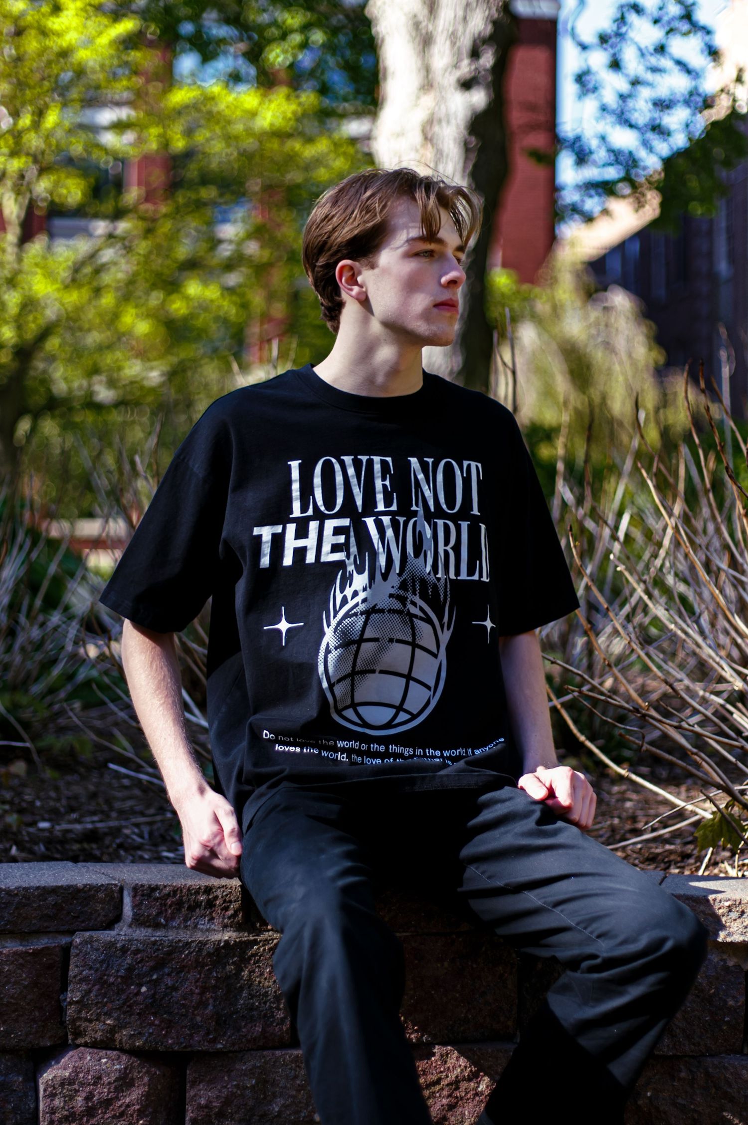 Young man seated on stone wall wearing black Love Not The World oversized graphic tee outdoors.